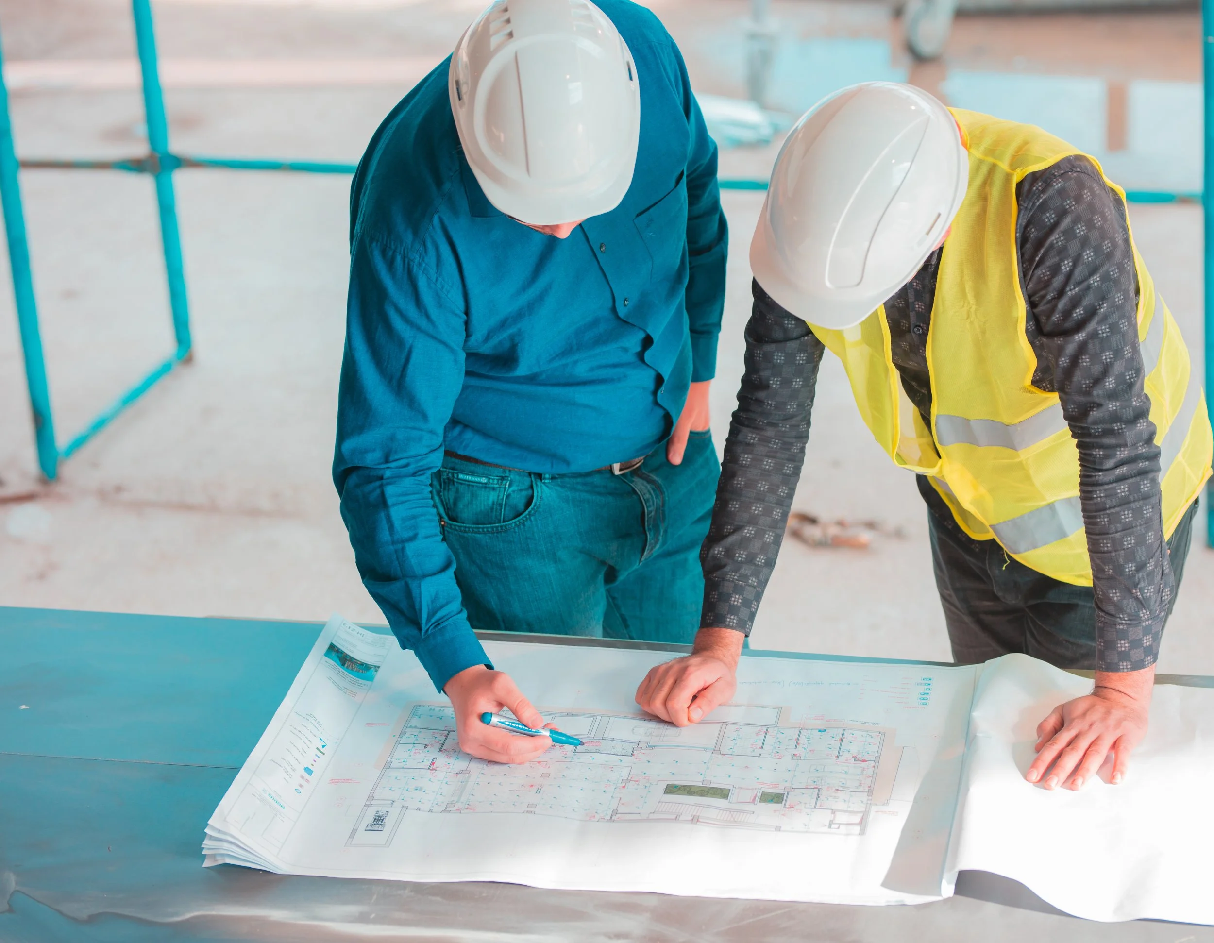 Two construction workers in safety helmets and vests examining architectural plans on a table at a construction site.