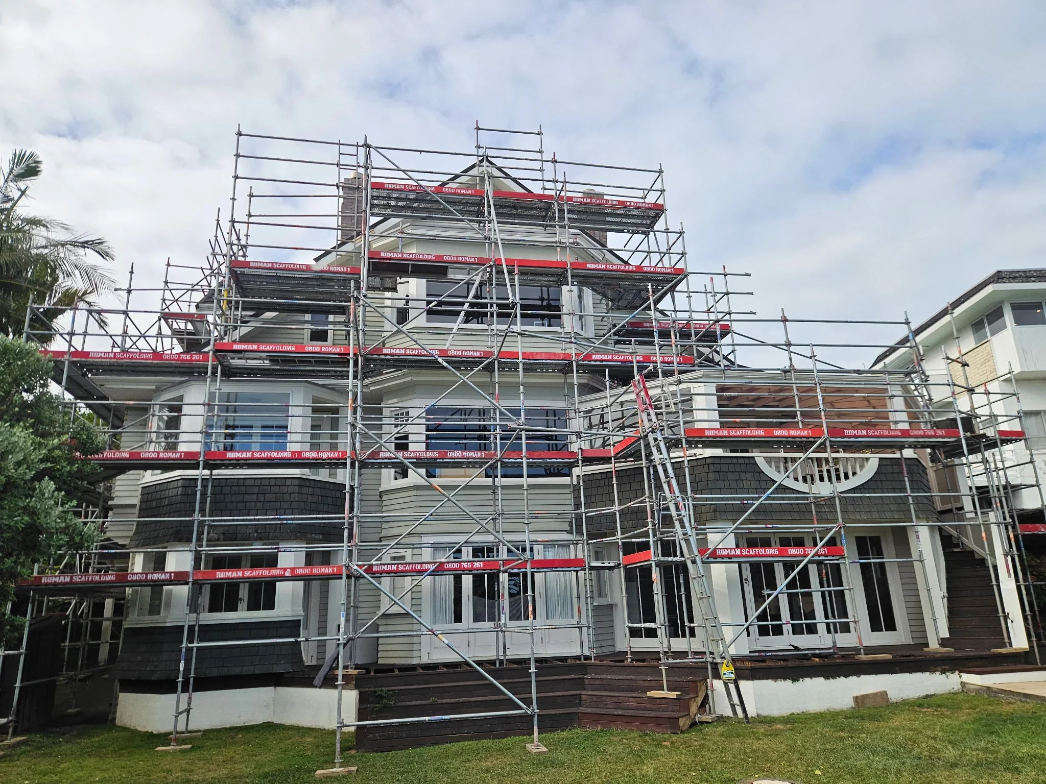 A house under renovation with scaffolding around it, including multiple levels with red and white signage for Roman Scaffolding, under a partly cloudy sky.
