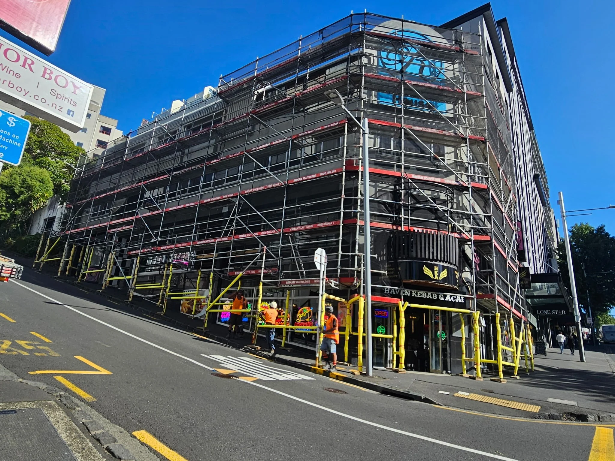 A multi-story building under construction covered with scaffolding, with construction workers at the base, and a clear blue sky in the background.