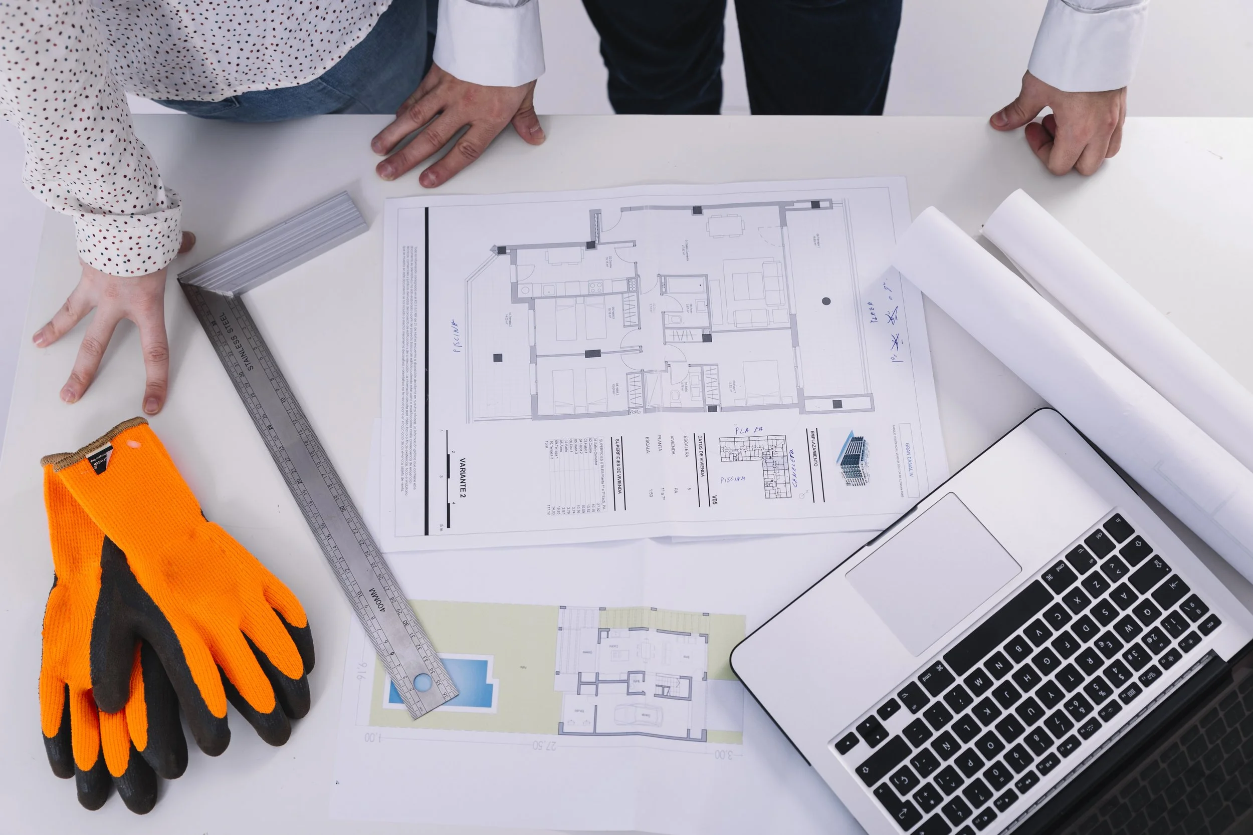 Blueprints for a building on a desk with a laptop, work gloves, a ruler, and orange safety gloves.