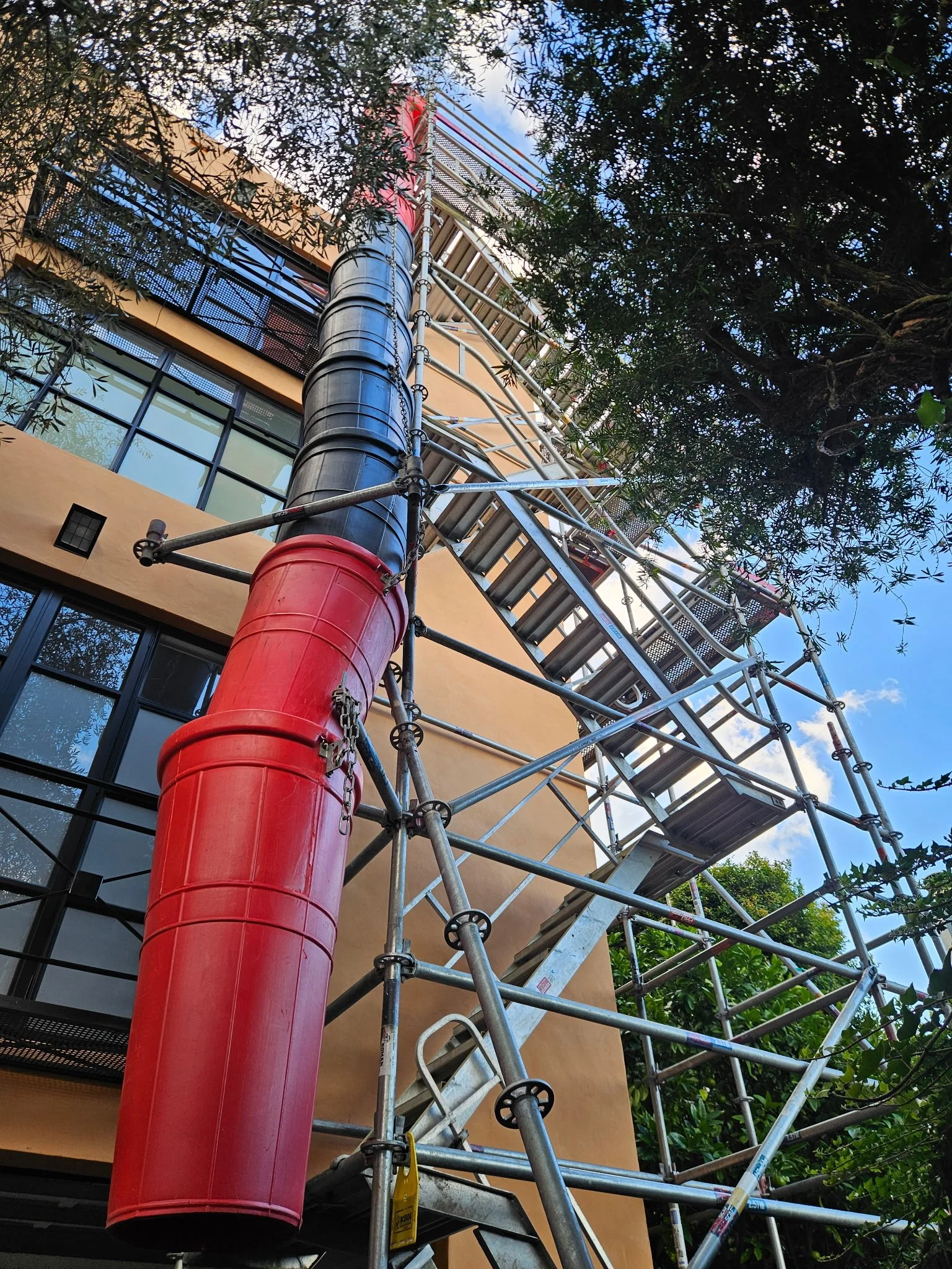 View of a building under construction with scaffolding and orange walls, surrounded by trees, with a blue sky in the background.