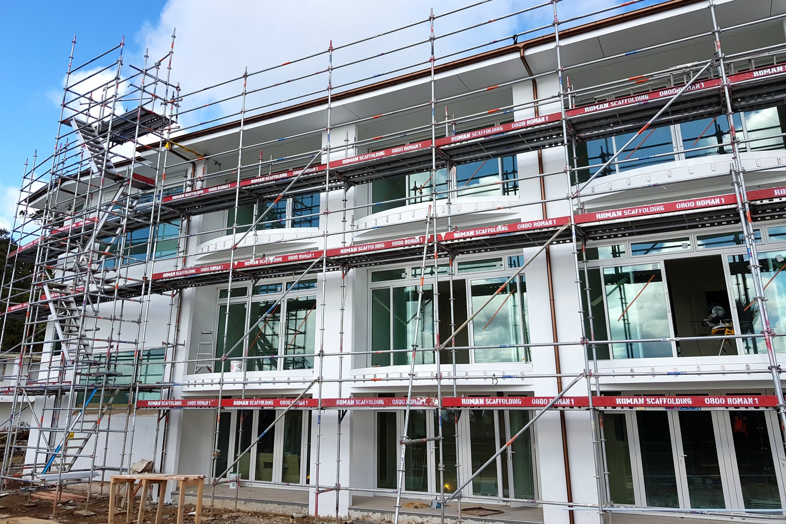 Scaffolding surrounds a modern building under construction, with large glass windows and multiple floors, against a partly cloudy sky.