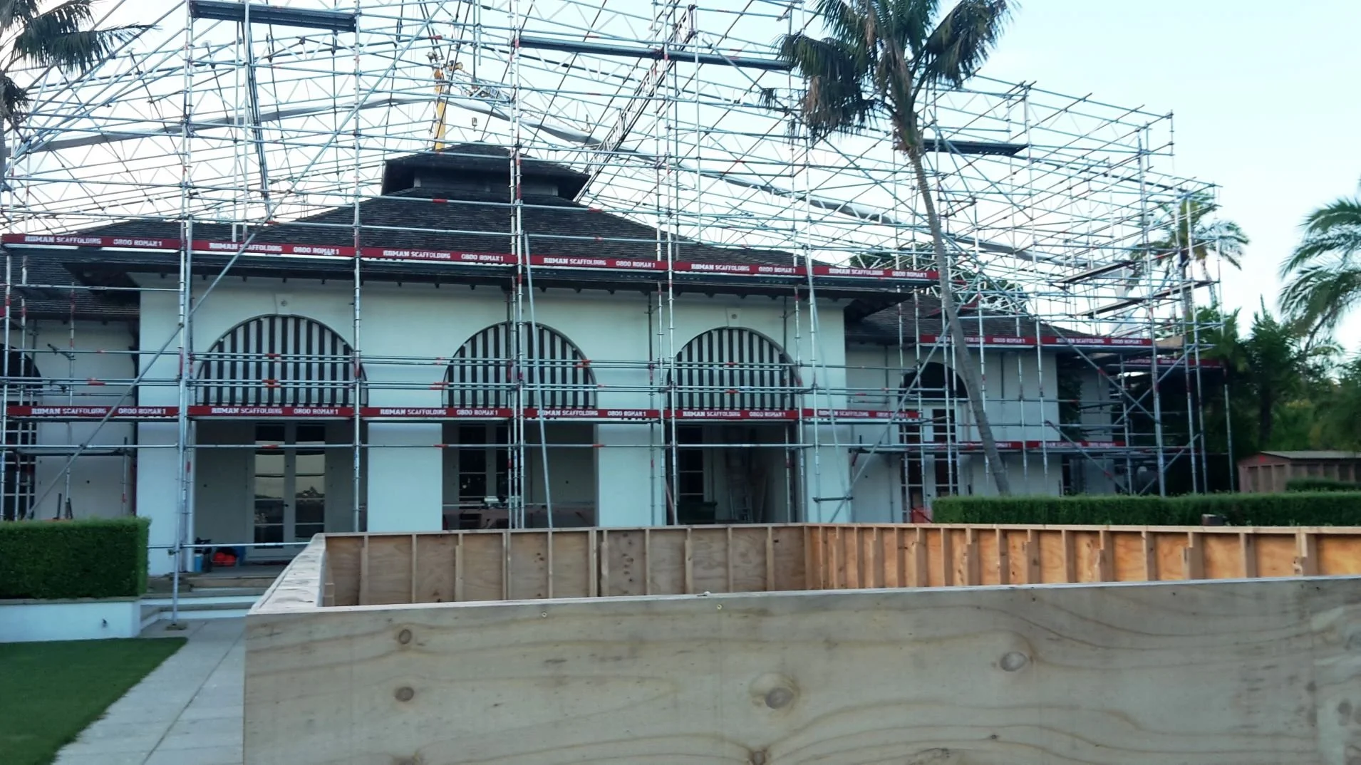 A house under construction with scaffolding around it, front yard with green lawn, and palm trees in the background.