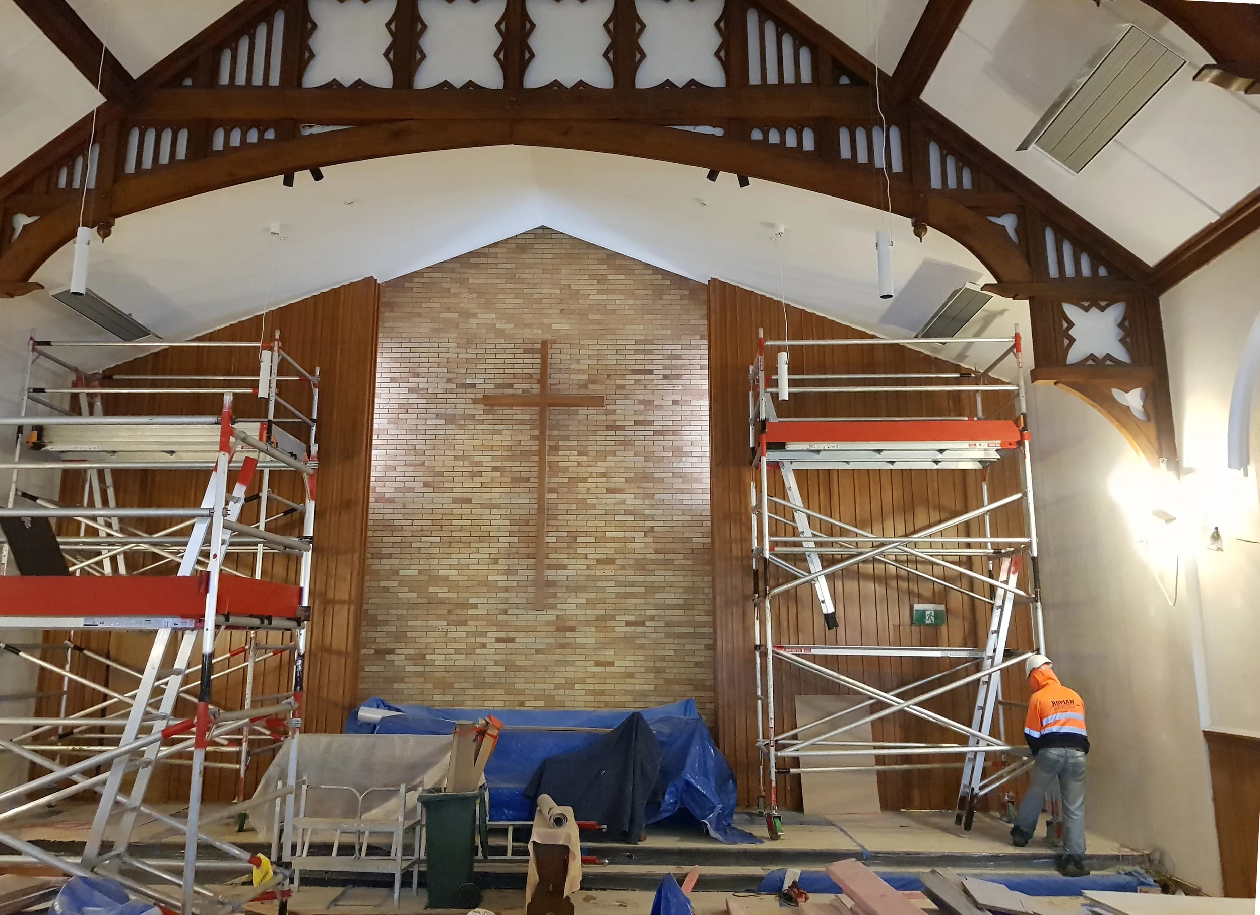 The interior of a church or chapel under renovation, with scaffolding set up in front of the altar, which has a large wooden cross on a brick wall backdrop. Workers are present, with one visible at the bottom right wearing an orange safety vest.