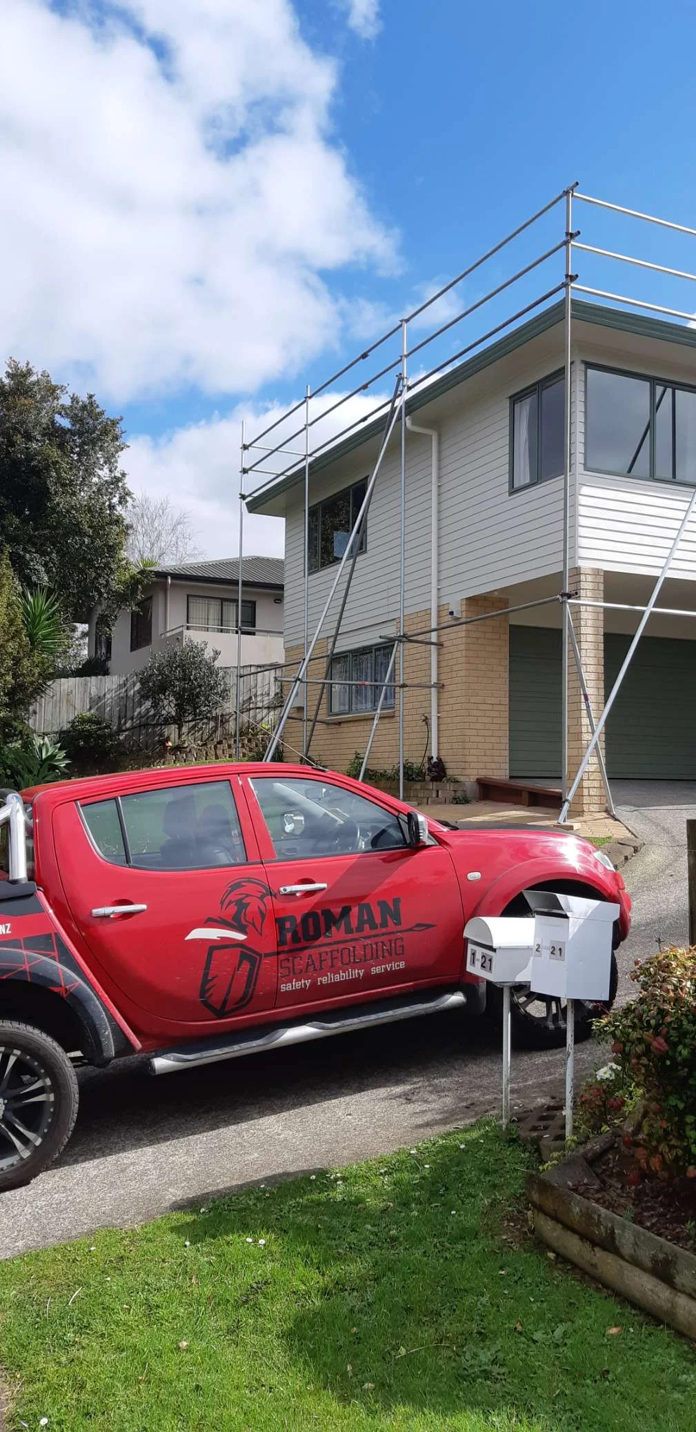 A red work truck labeled 'Roman Scaffoldings' parked in front of a two-story house with scaffolding around its upper floor, under a partly cloudy sky.