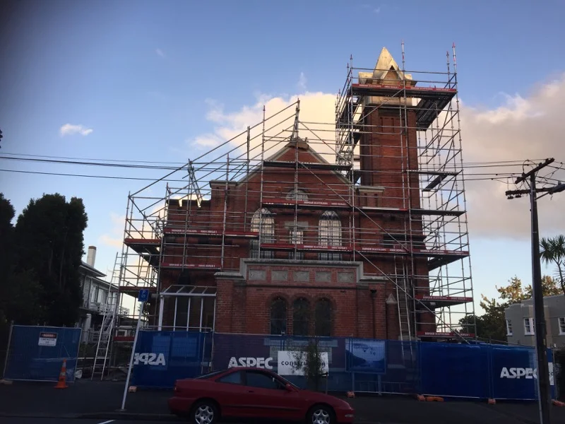 A brick building under construction with scaffolding surrounding it and a blue construction fence in the foreground.