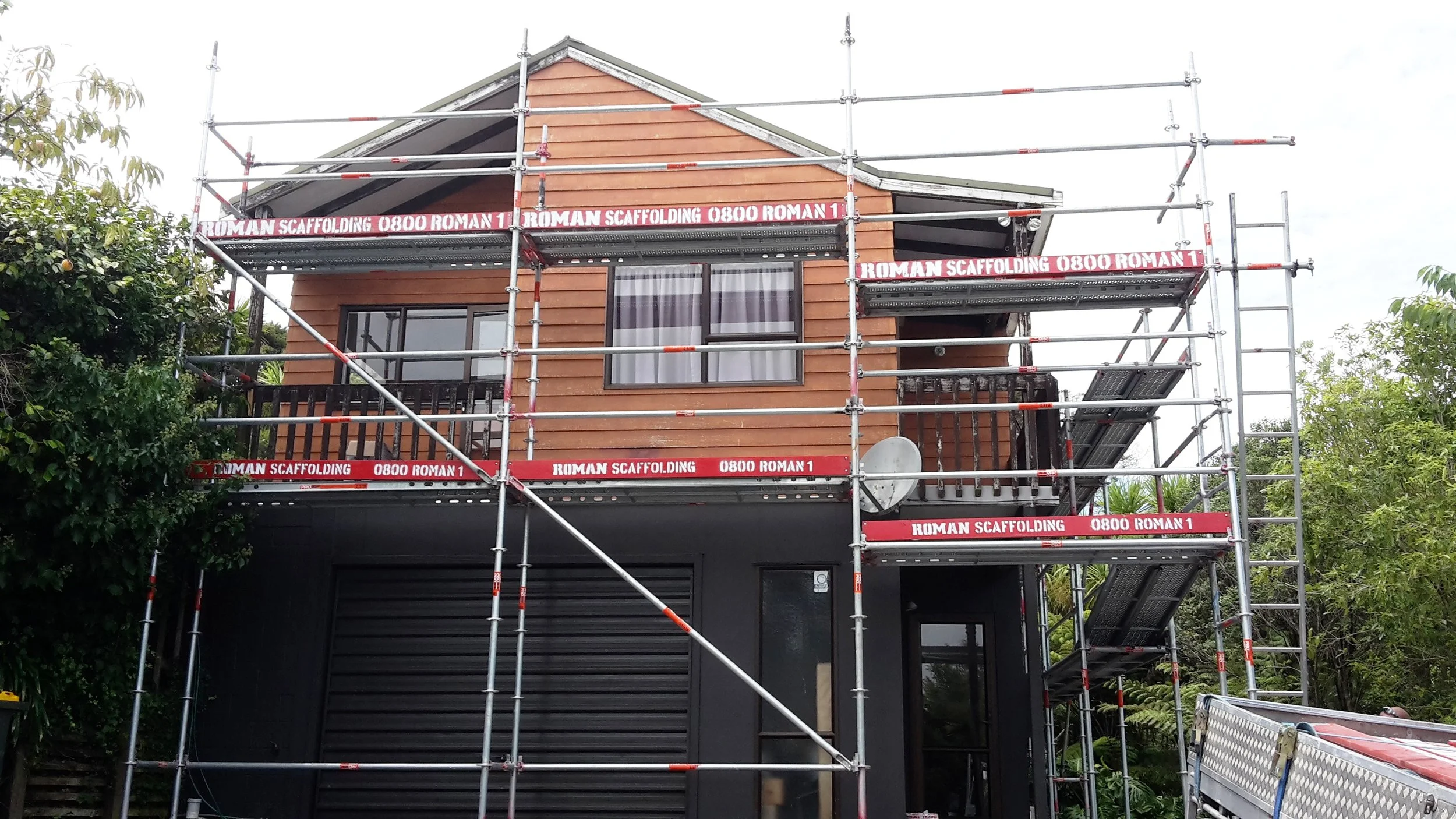A two-story house under construction with scaffolding around it. The house has wooden siding, two windows, and a satellite dish. The scaffolding is labeled 'Roman Scaffolding 0800 Roman 1'.