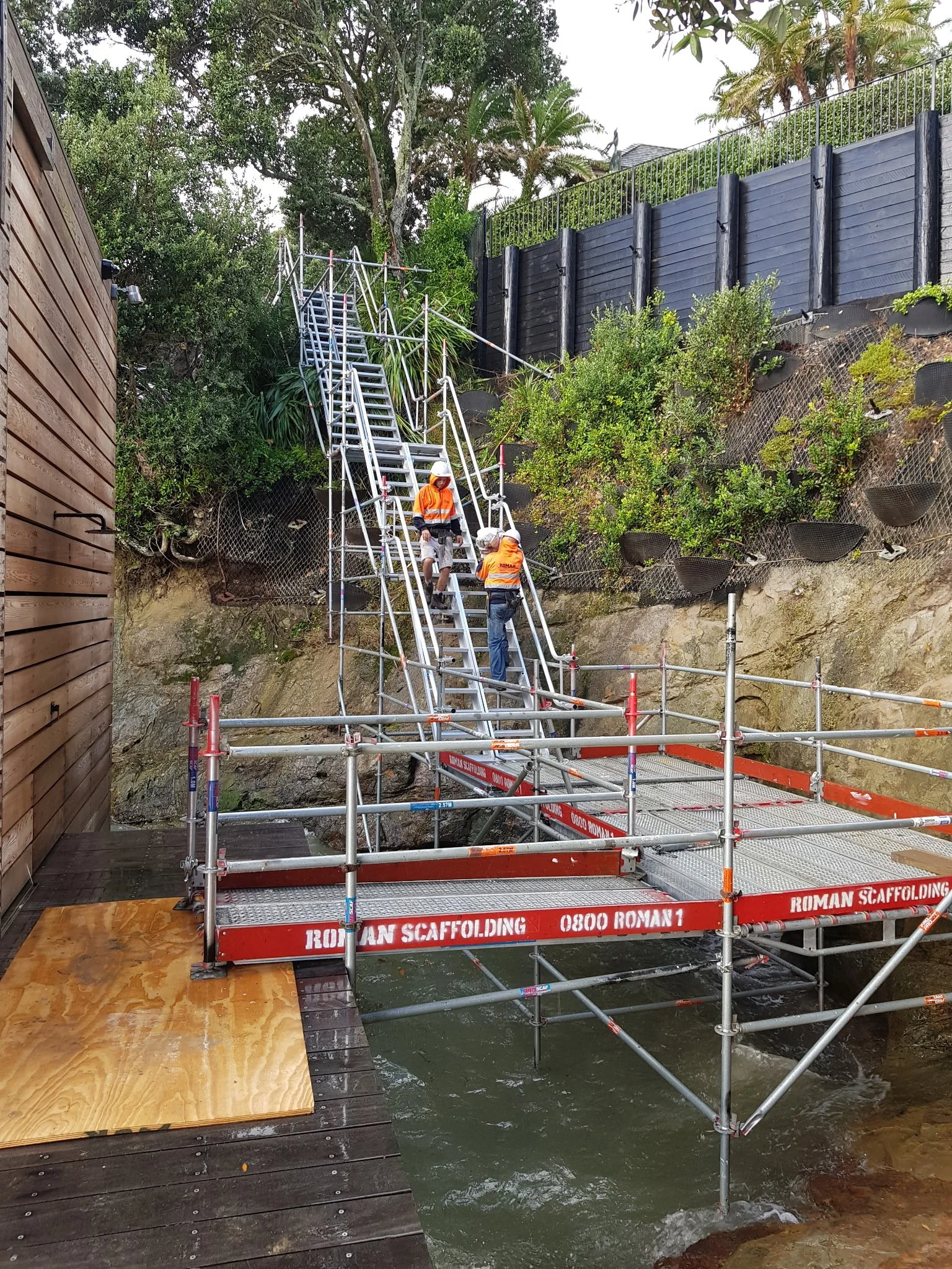 Construction workers installing scaffolding on a small body of water, with a wooden platform, rocks, plants, and a black fence on a hillside in the background.