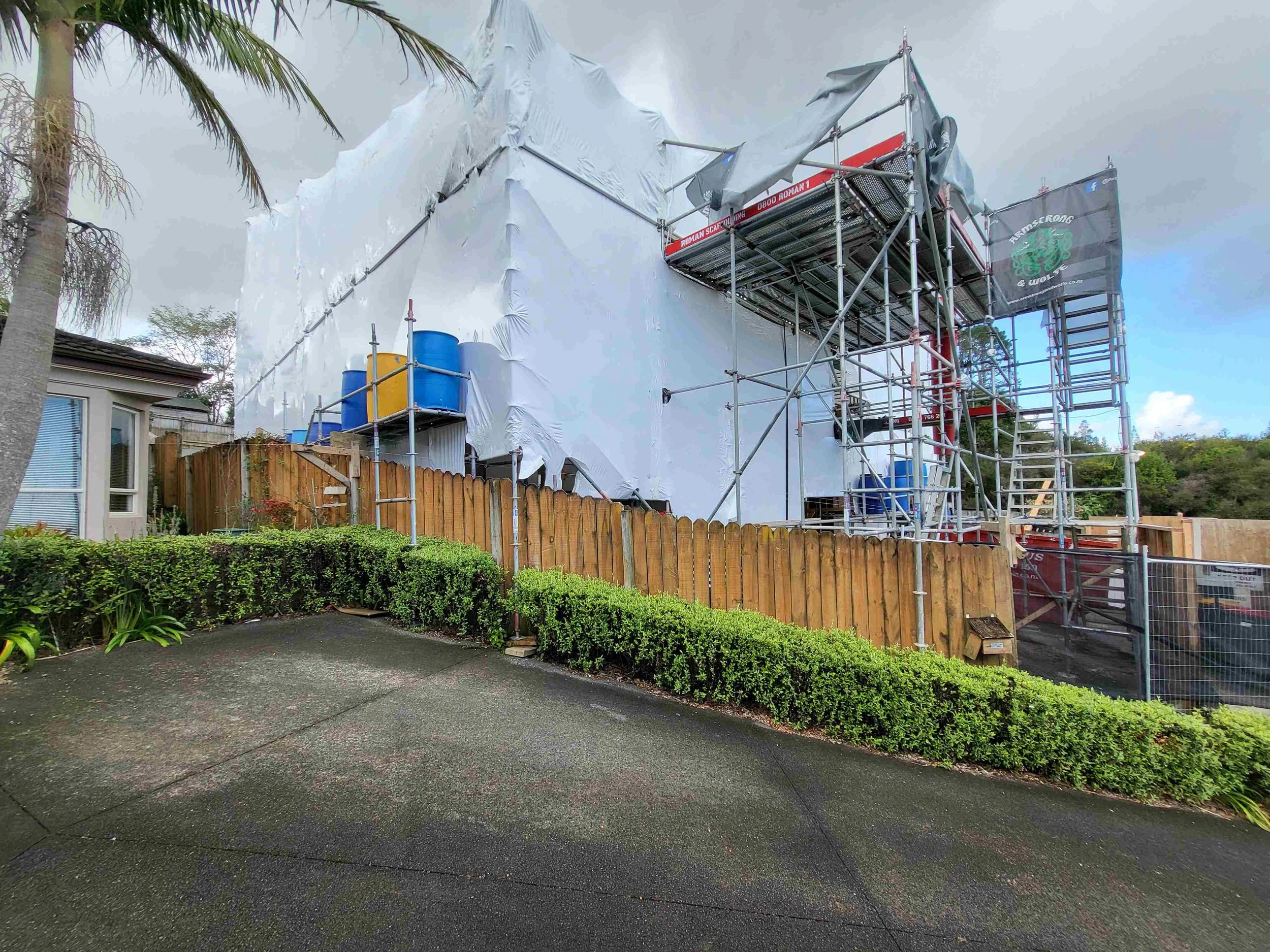 Construction scaffolding set up around the side of a residential house, with a large white tarp covering part of the building, in a suburban neighborhood with trees and a cloudy sky.