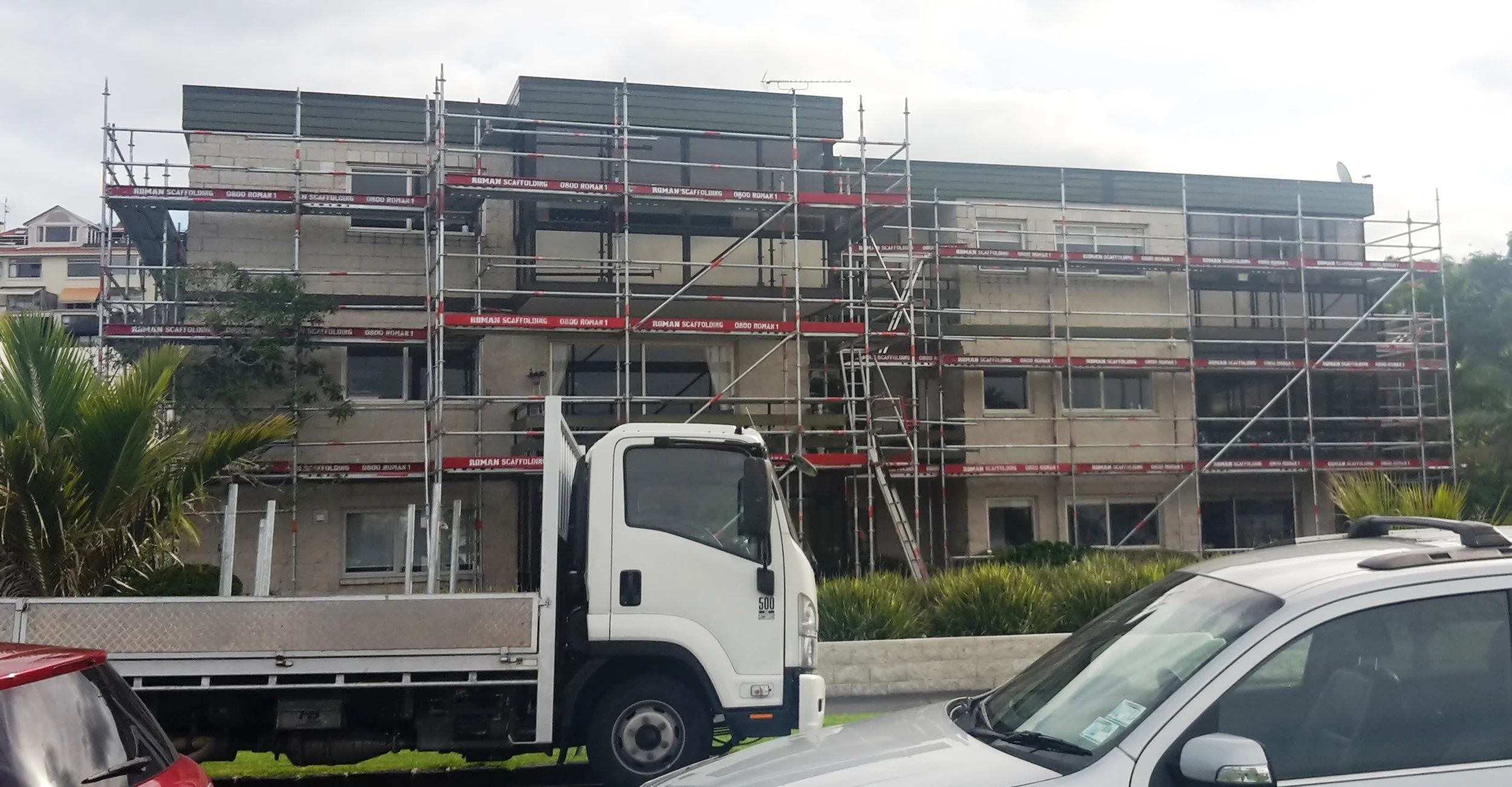 Construction site of a multi-story building with scaffolding surrounding it, and a white truck in the foreground.