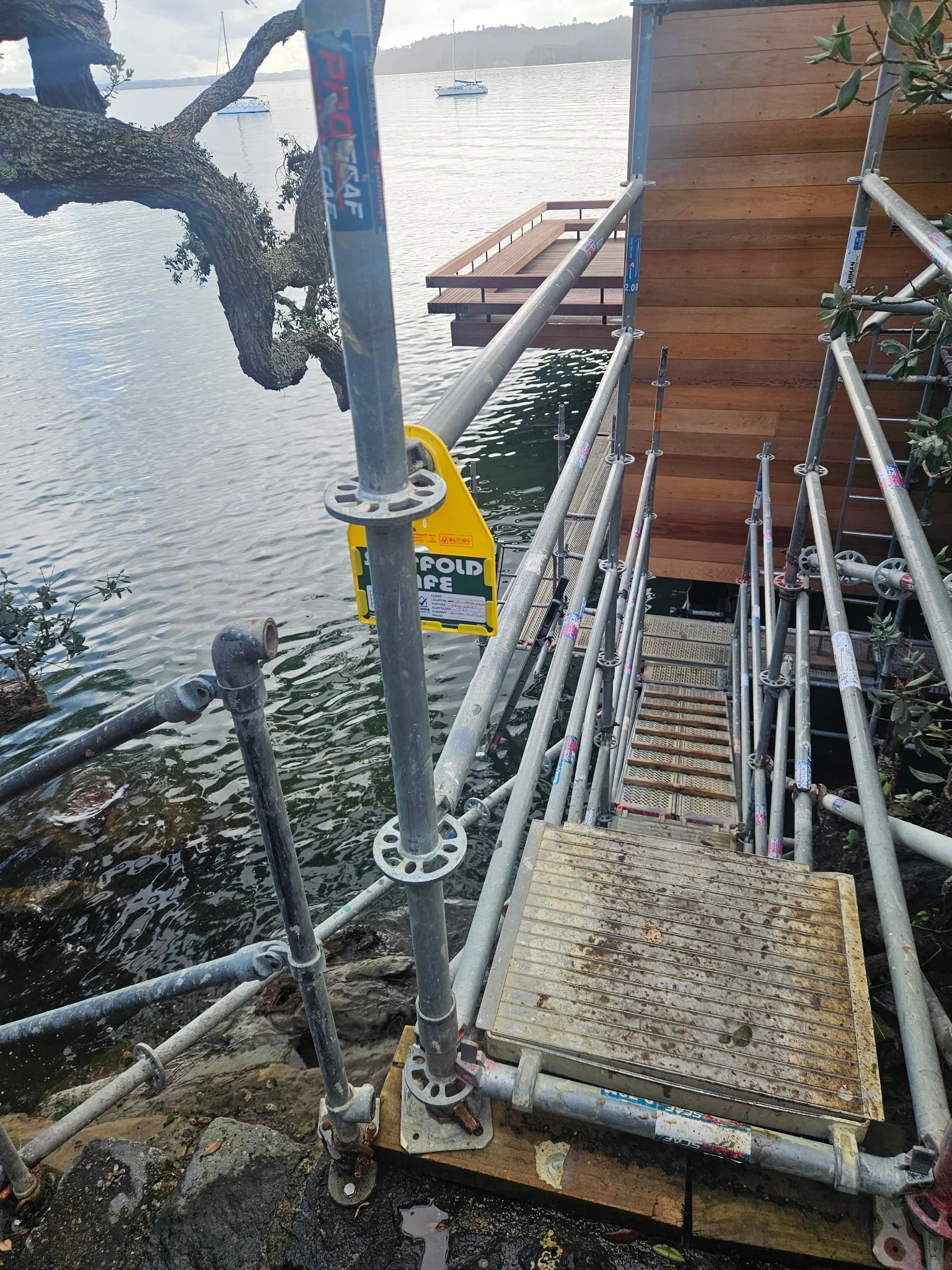 A construction scaffold set up next to a wooden deck over a body of water with boats and trees in the background.