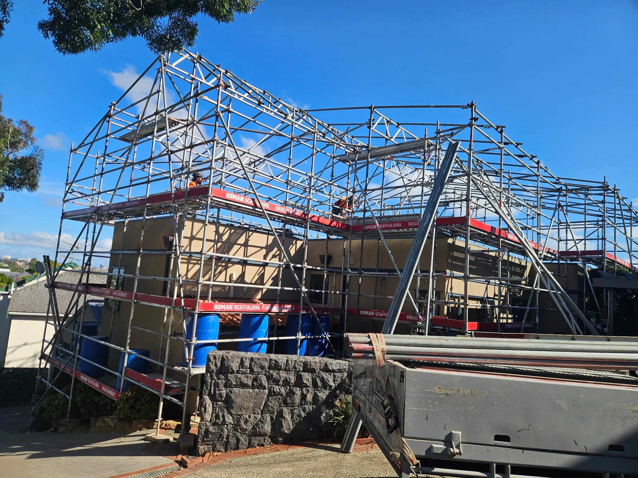 A building under construction with metal scaffolding, two workers in safety gear, and a clear blue sky.