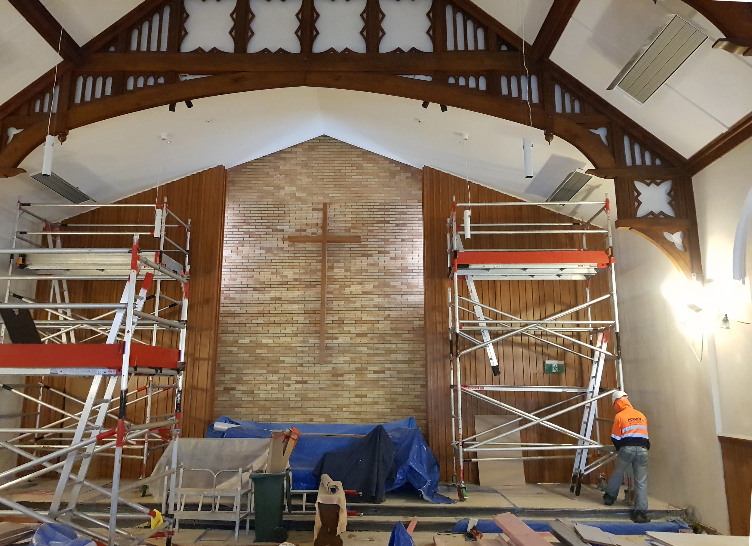 Inside a church or chapel, with wooden ceiling beams and a brick wall with a cross. There are scaffolding and construction workers indicating ongoing renovation or construction work.