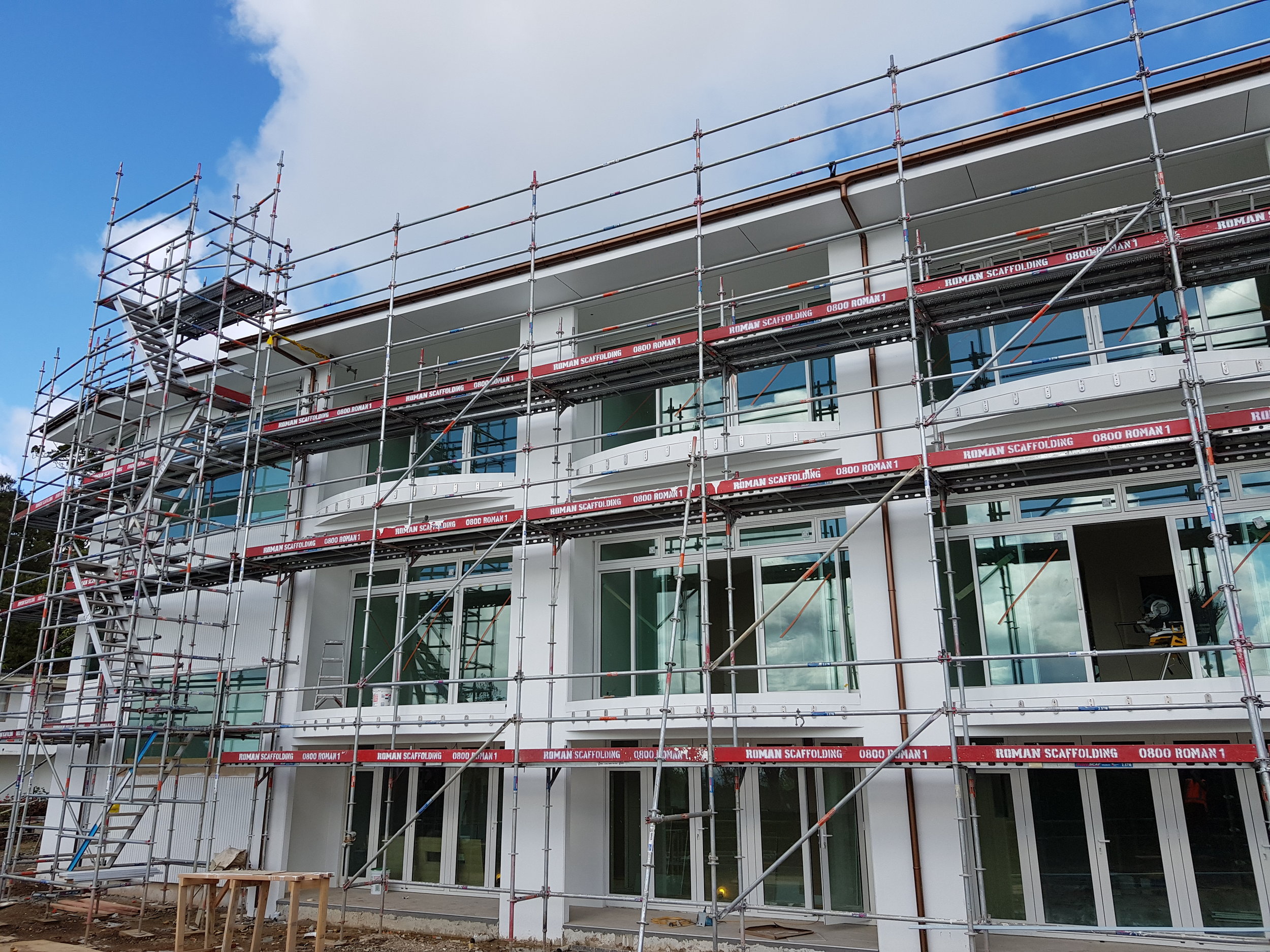 Construction scaffolding surrounds a modern building with large glass windows, during daytime with partly cloudy sky.
