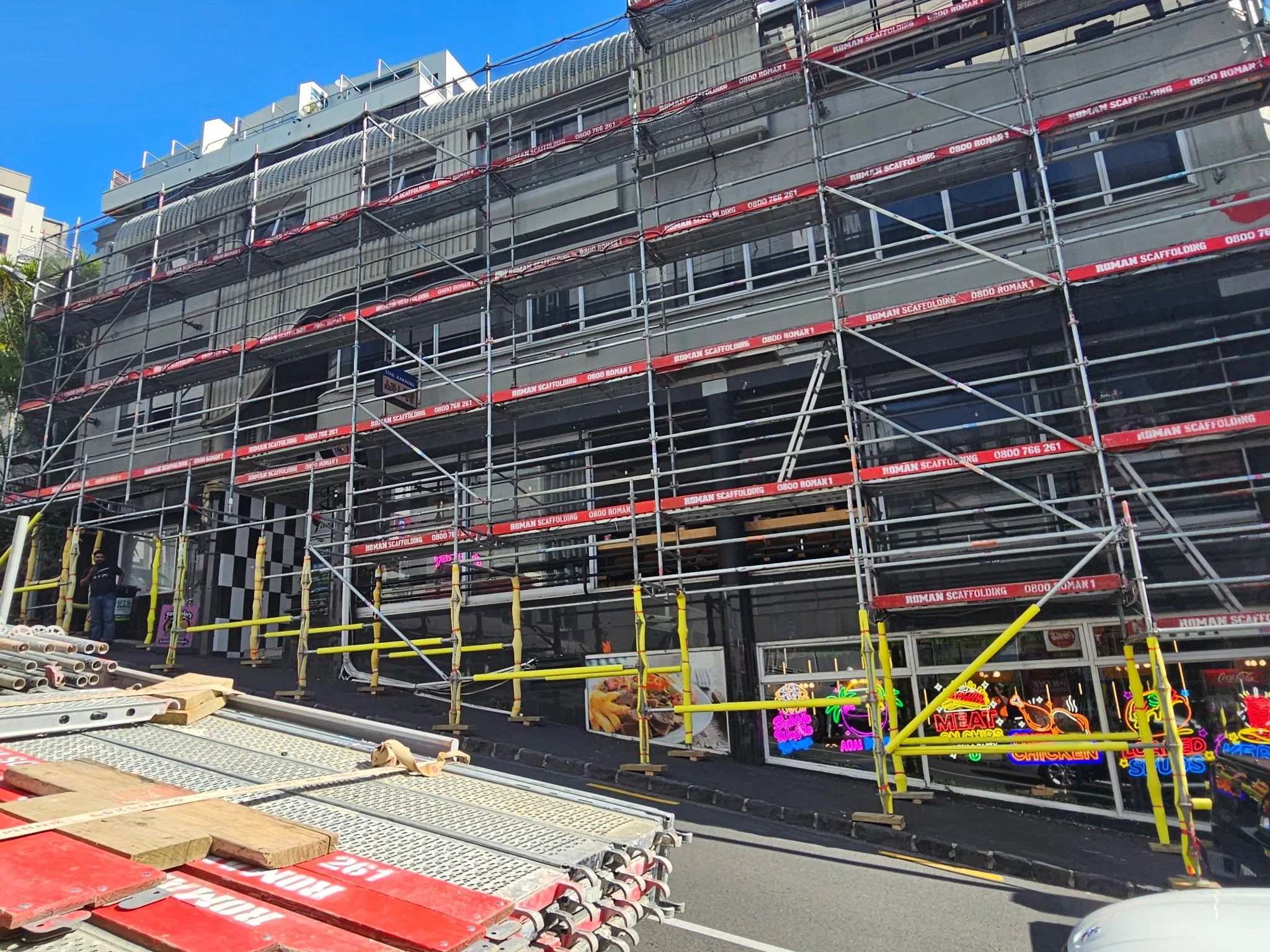 Building under construction with scaffolding surrounding it, neon signs at the bottom advertise food and drink options, and construction materials are visible in the foreground.