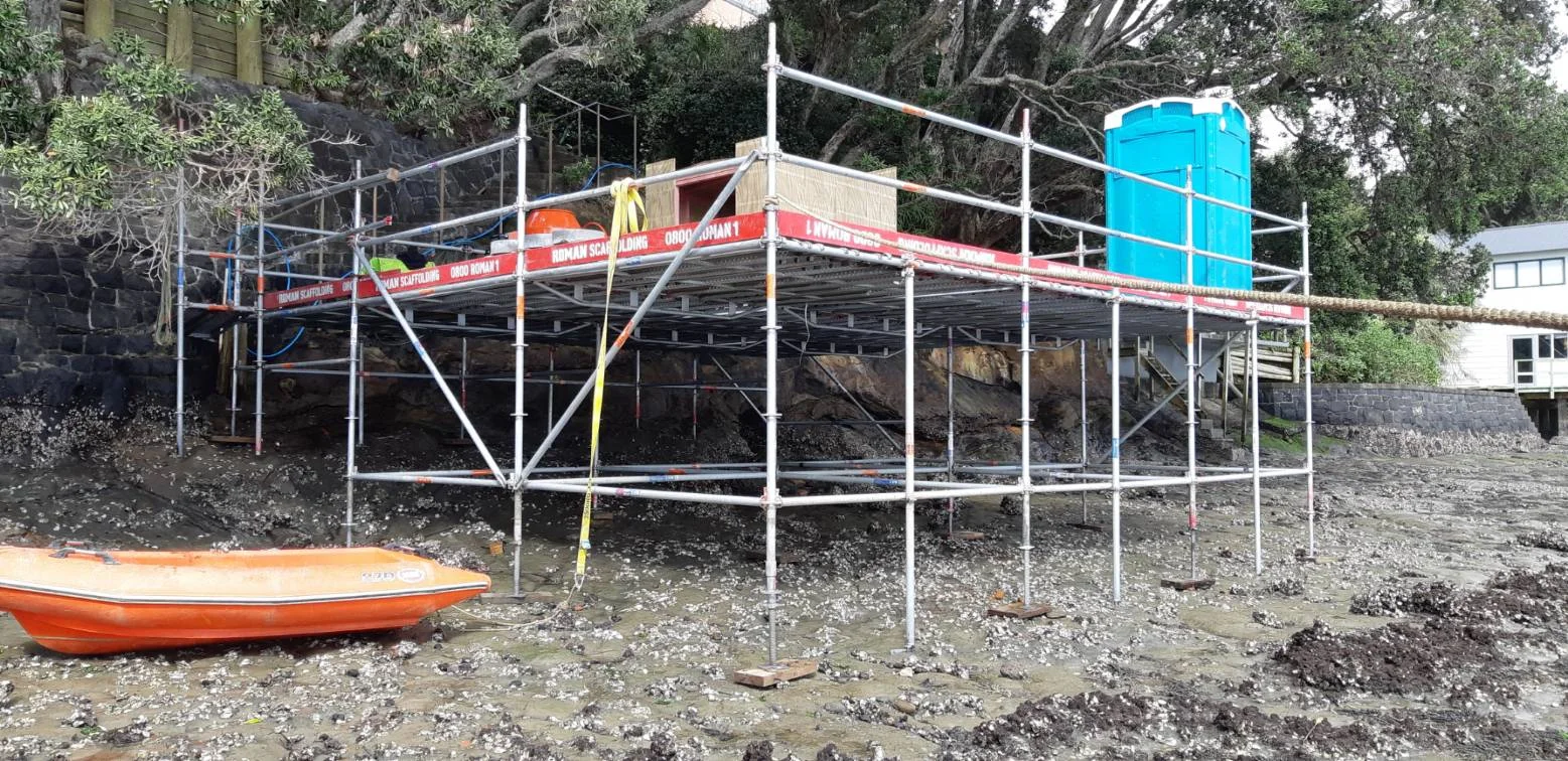 Construction scaffolding set up on a rocky beach with a portable toilet and a small boat nearby.