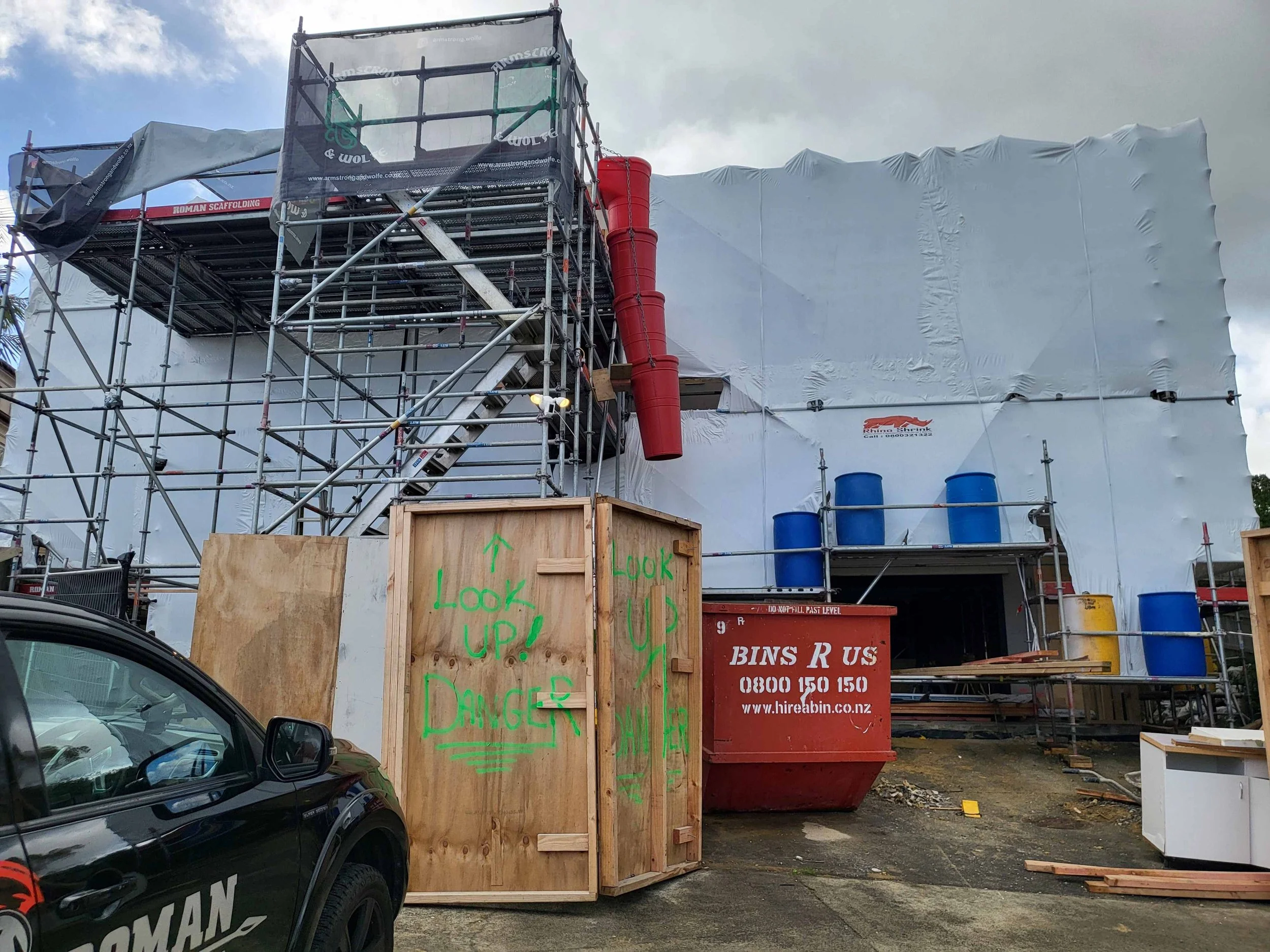 Construction site with scaffolding, wrapped structures, and a red trash bin. A wooden panel with green handwritten signs and a part of a black car are visible in the foreground.