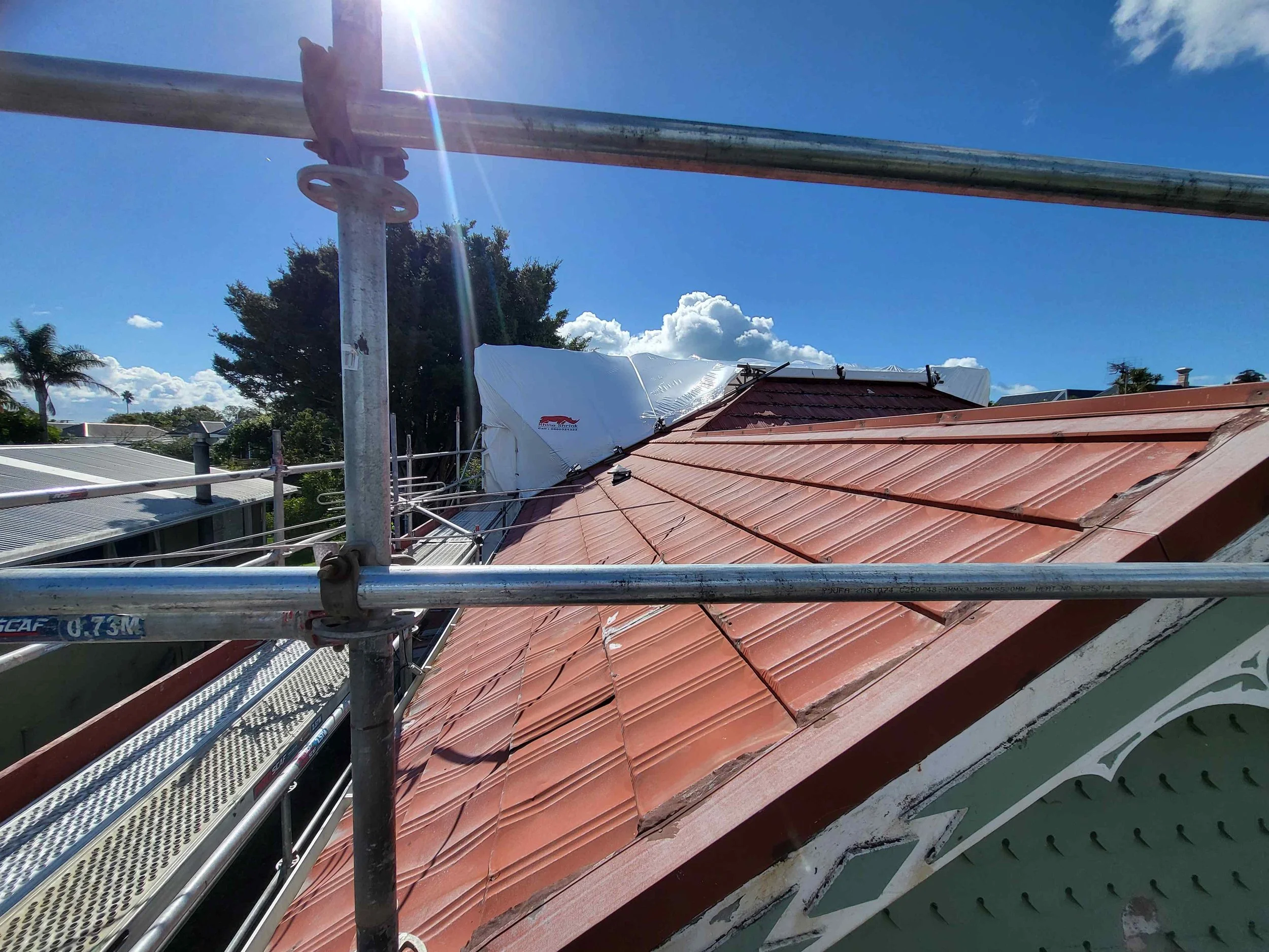 Photo of a house roof with red tiles under blue sky, scaffolding and sun glare visible.