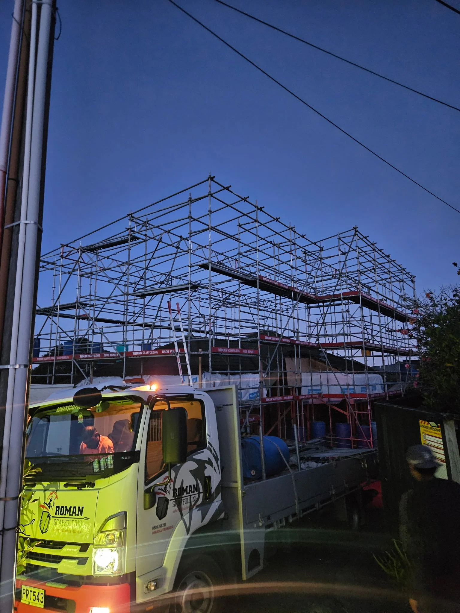 A work truck with scaffolding equipment on the back is parked in front of a building under construction at dusk. The truck has a logo that says 'ROMAN SCAFFOLDING' and is illuminated by a streetlight.