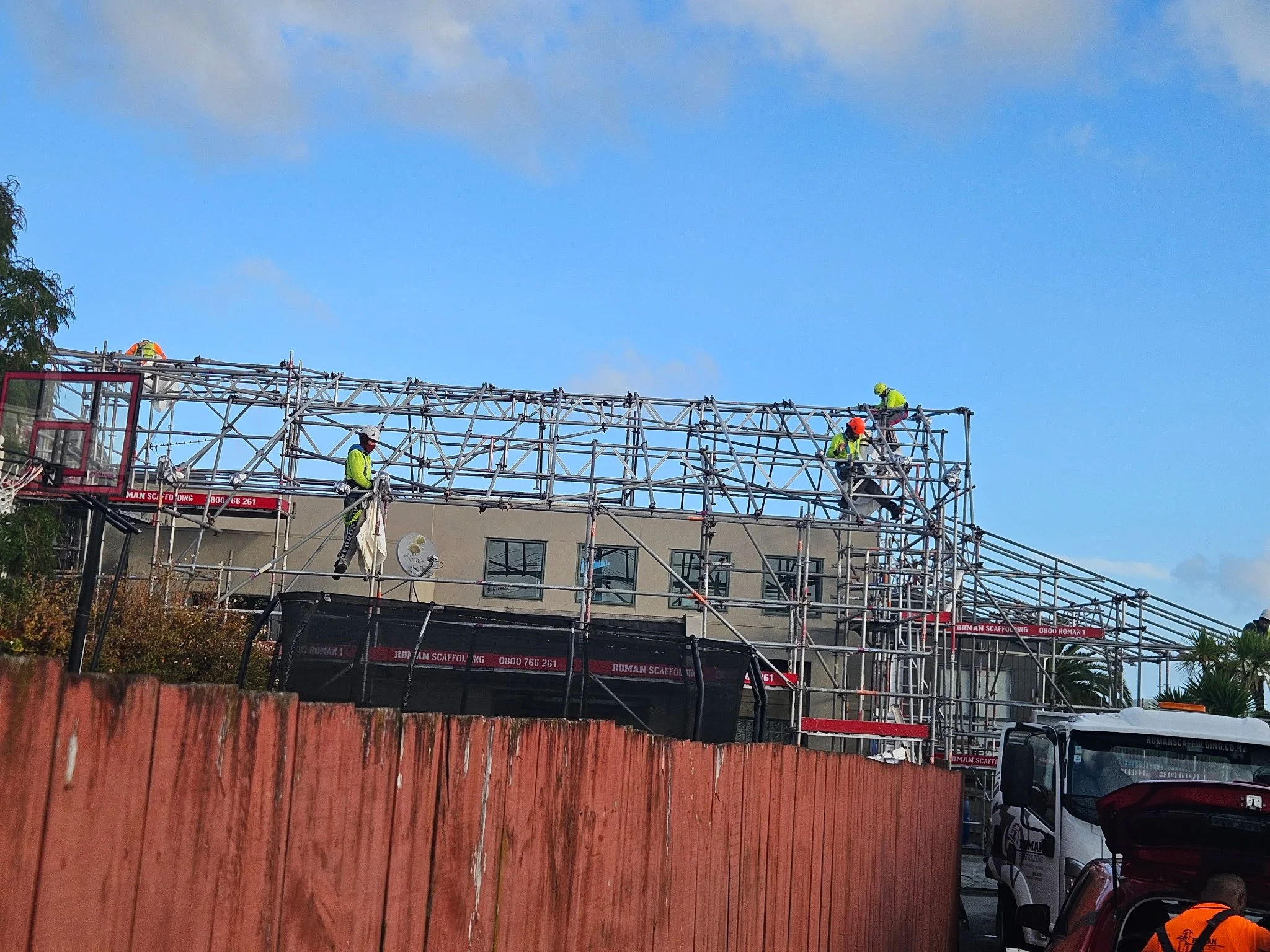 Construction workers assembling a metal scaffolding structure outdoors, with a building and trees in the background, under a partly cloudy sky.