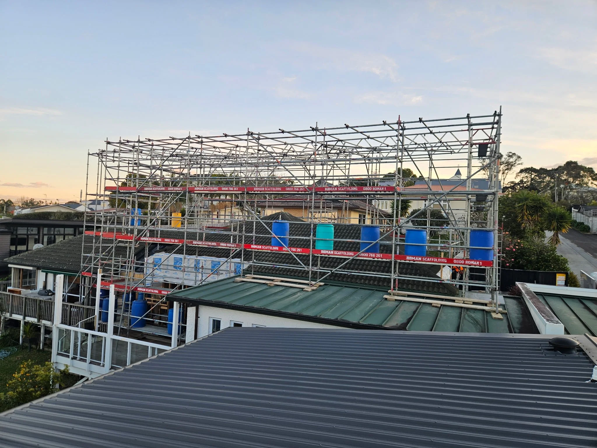 Scaffolding structure around a house under construction in a neighborhood with trees and rooftops visible at dusk.