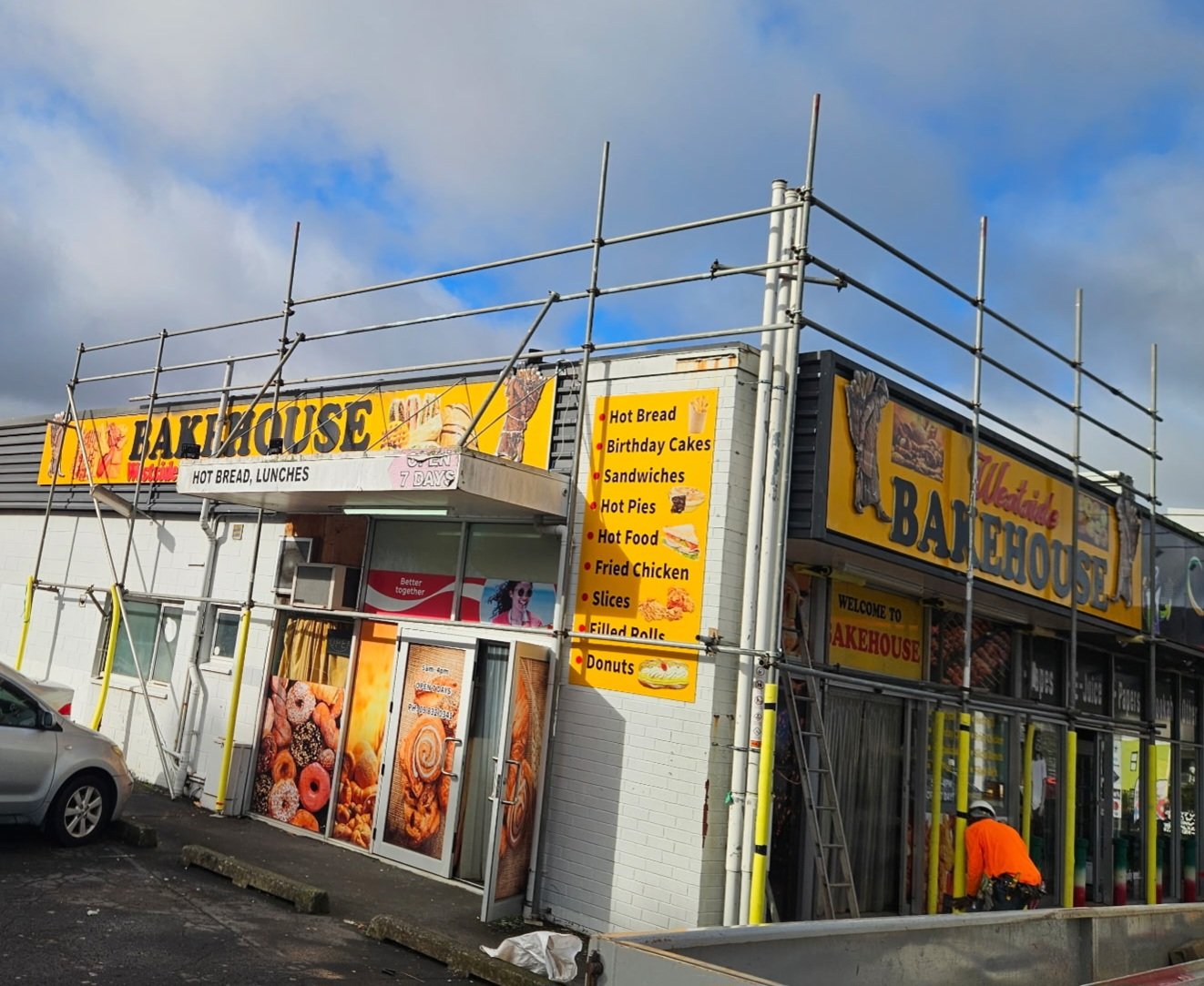 Exterior view of a fast-food restaurant called 'Bakehouse' under construction, with scaffolding around the building. The sign lists menu items such as hot bread, birthday cakes, sandwiches, hot pies, hot food, fried chicken, slices, filled rolls, and donuts. A man in orange clothing is working near the entrance, and a parked car is visible in the foreground.