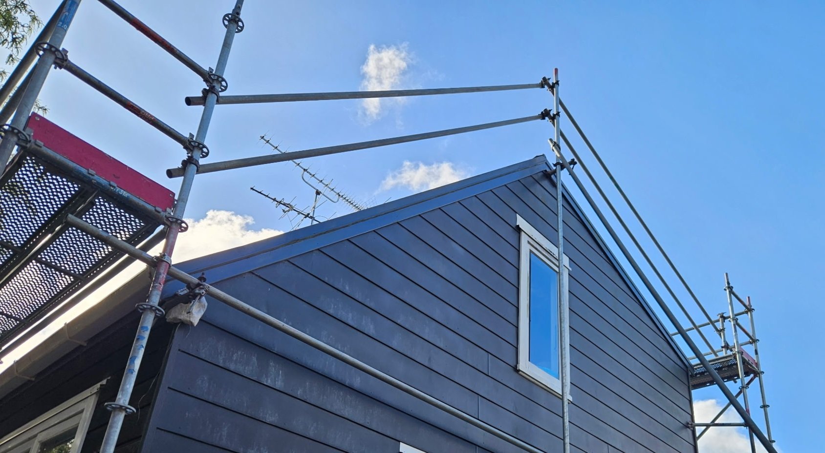 Construction scaffolding set up on a modern house with dark siding and a small window under a blue sky with clouds.
