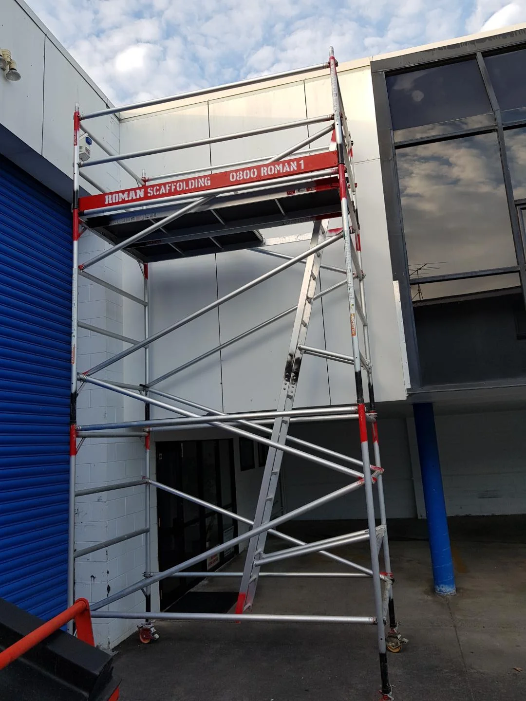 A metal scaffold tower labeled 'Roman Scaffolding' set up outside a building with a white wall and dark window, against a partly cloudy sky.