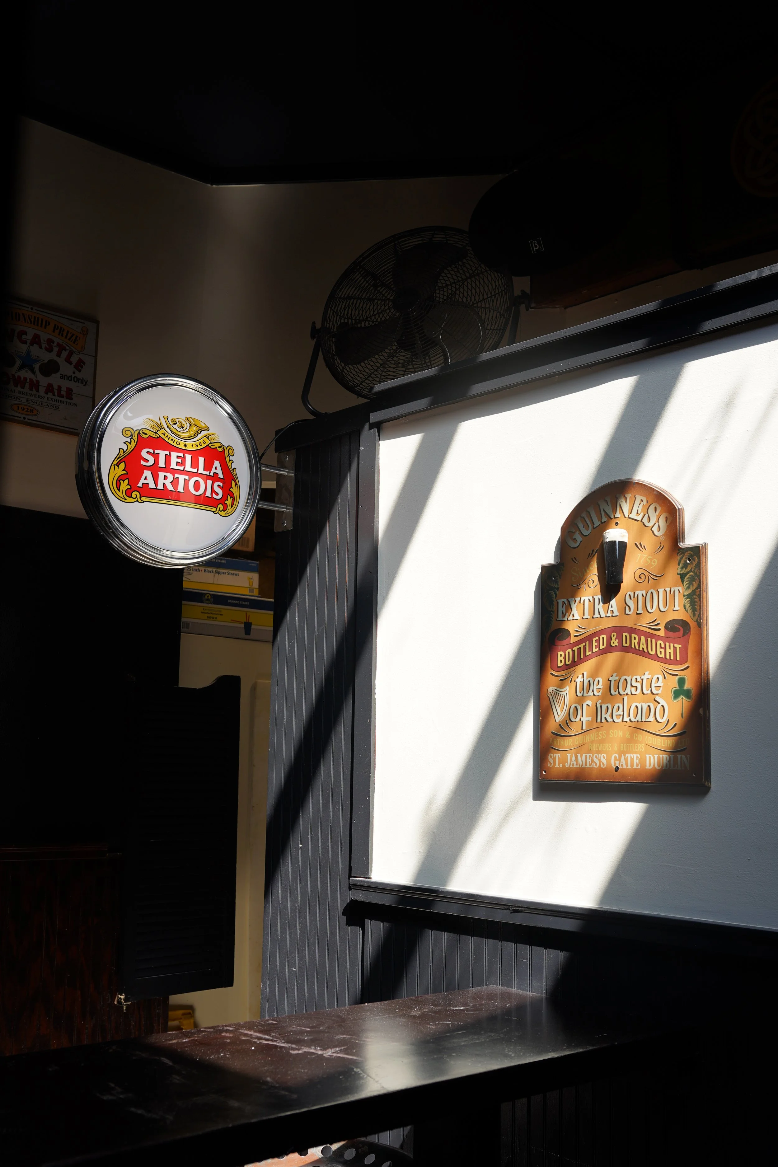 Interior of a pub or bar with a Stella Artois sign, a wind fan, and a Guinness sign on a white wall, with sunlight casting shadows.