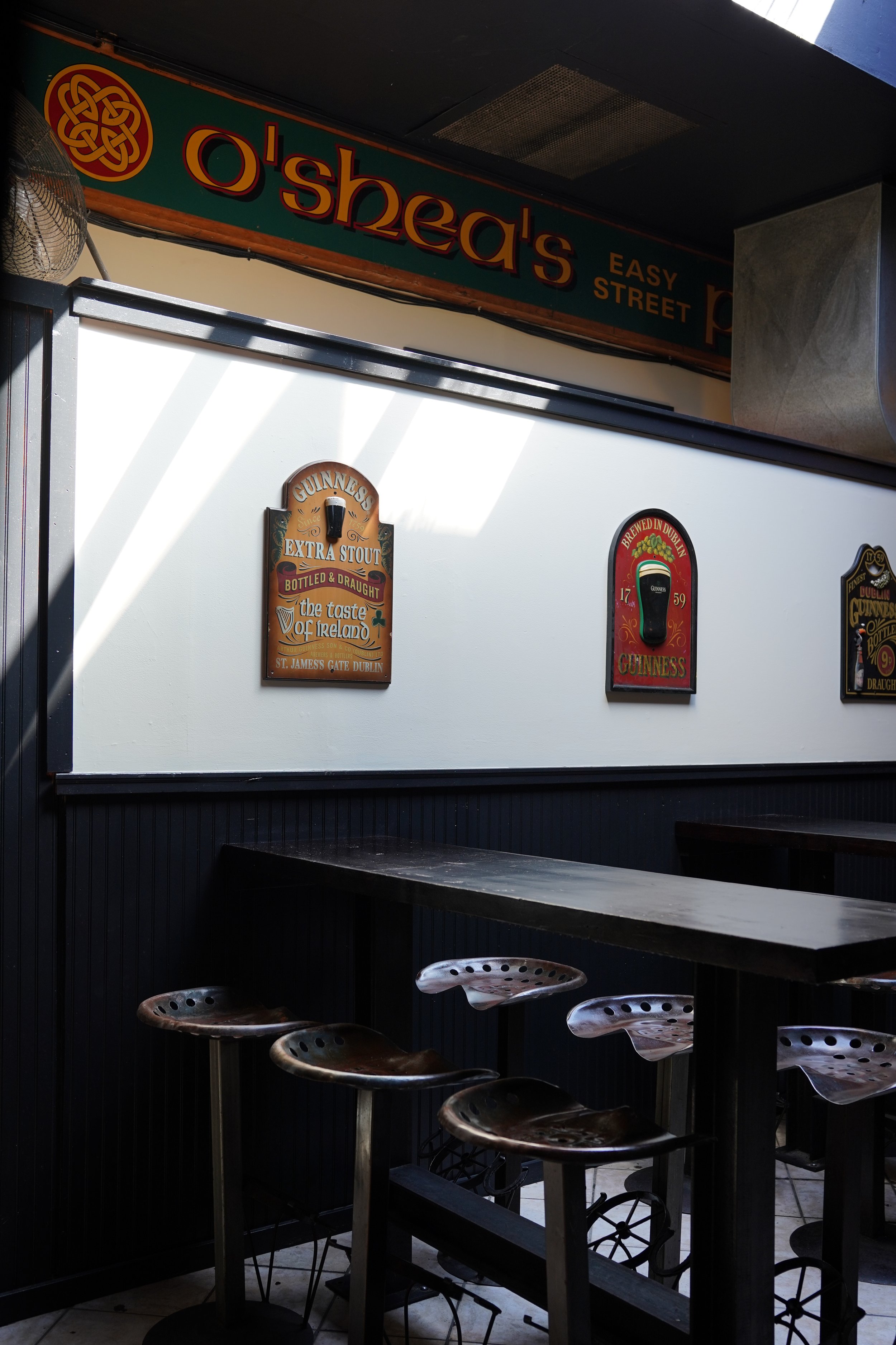 Interior of an Irish pub with advertisements for Guinness beer on the wall, a high table with metal stools, and a sign reading 'O'Shea's Easy Street'.