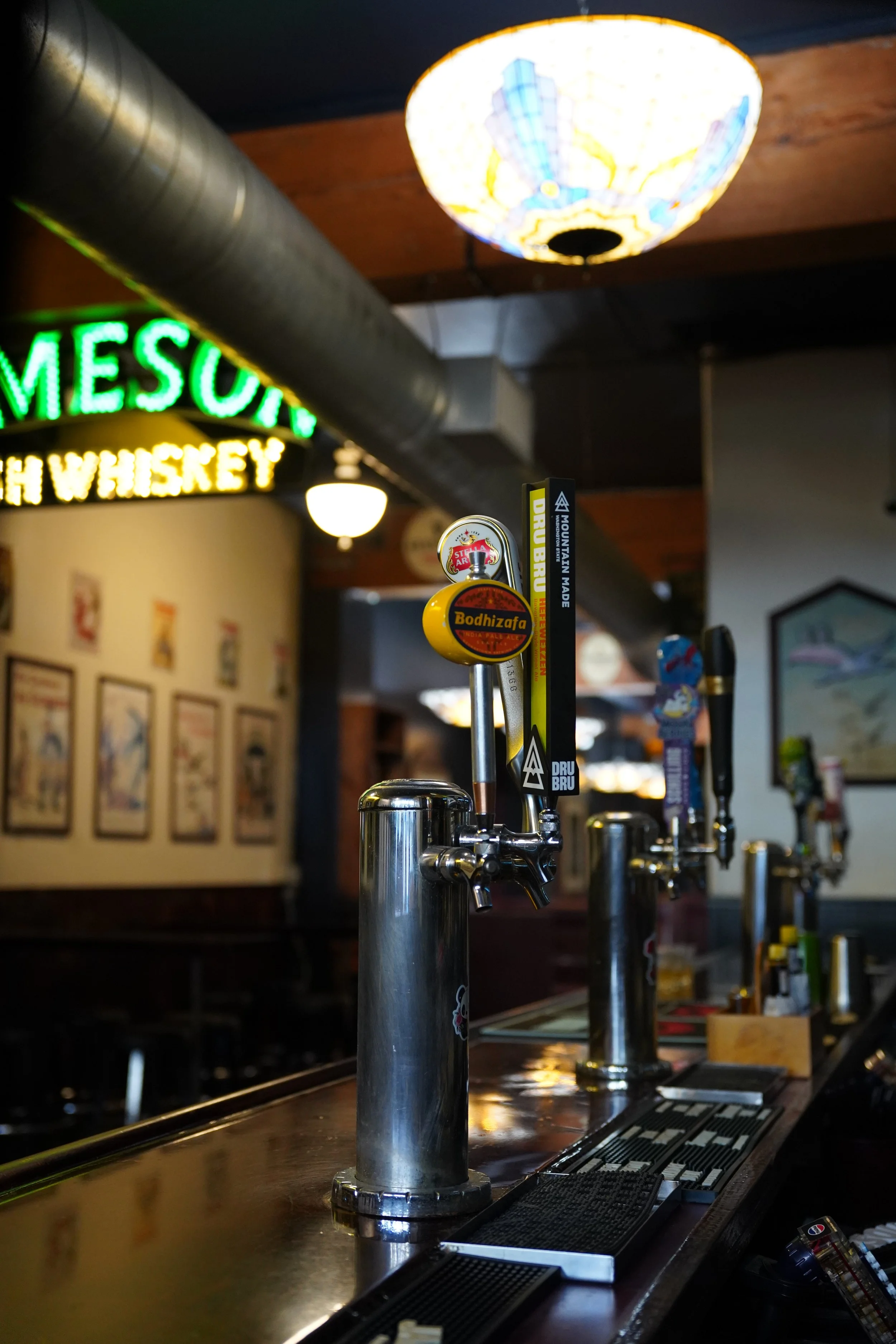 Multiple beer taps at a bar with colorful drink decals and a neon sign in the background.