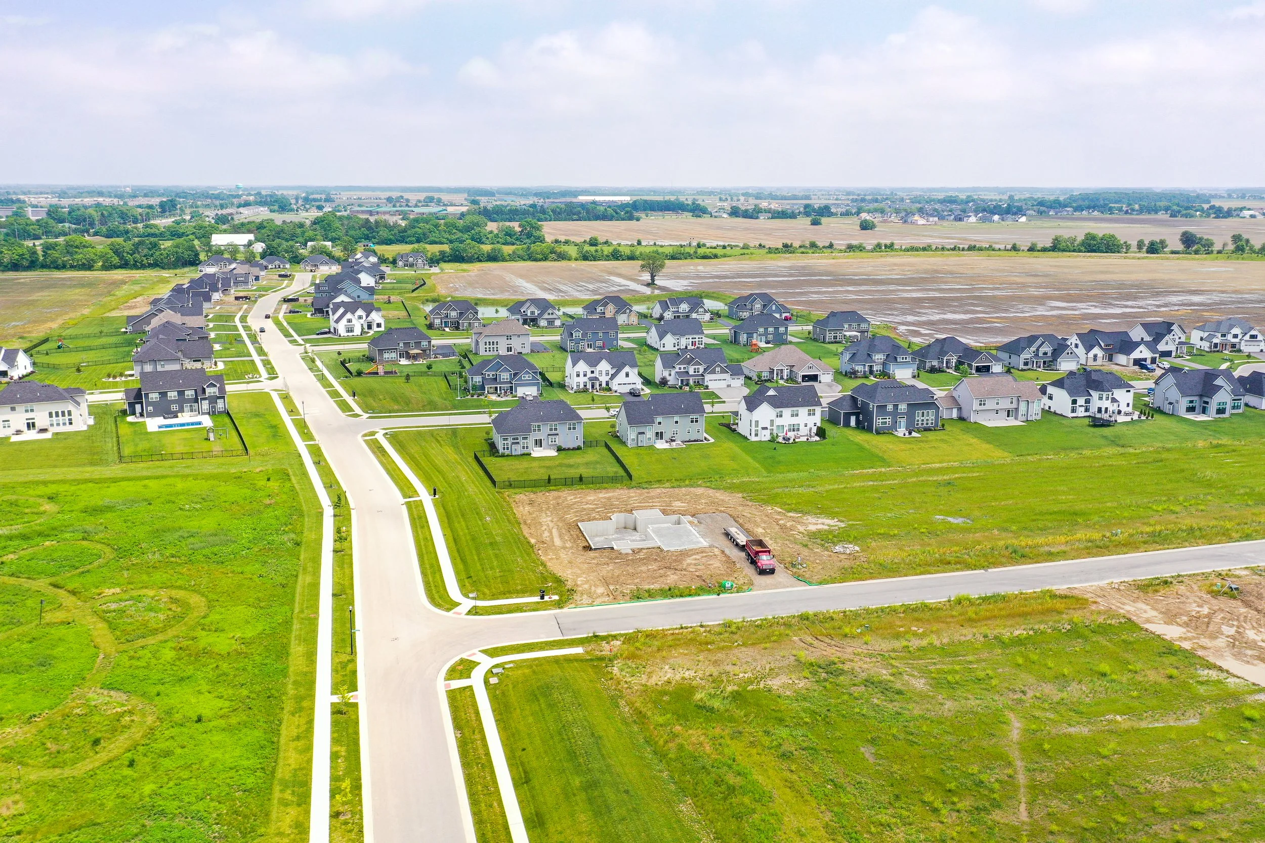 An aerial view of a suburban neighborhood with houses, roads, and manicured lawns, with fields and open land in the background under a partly cloudy sky.