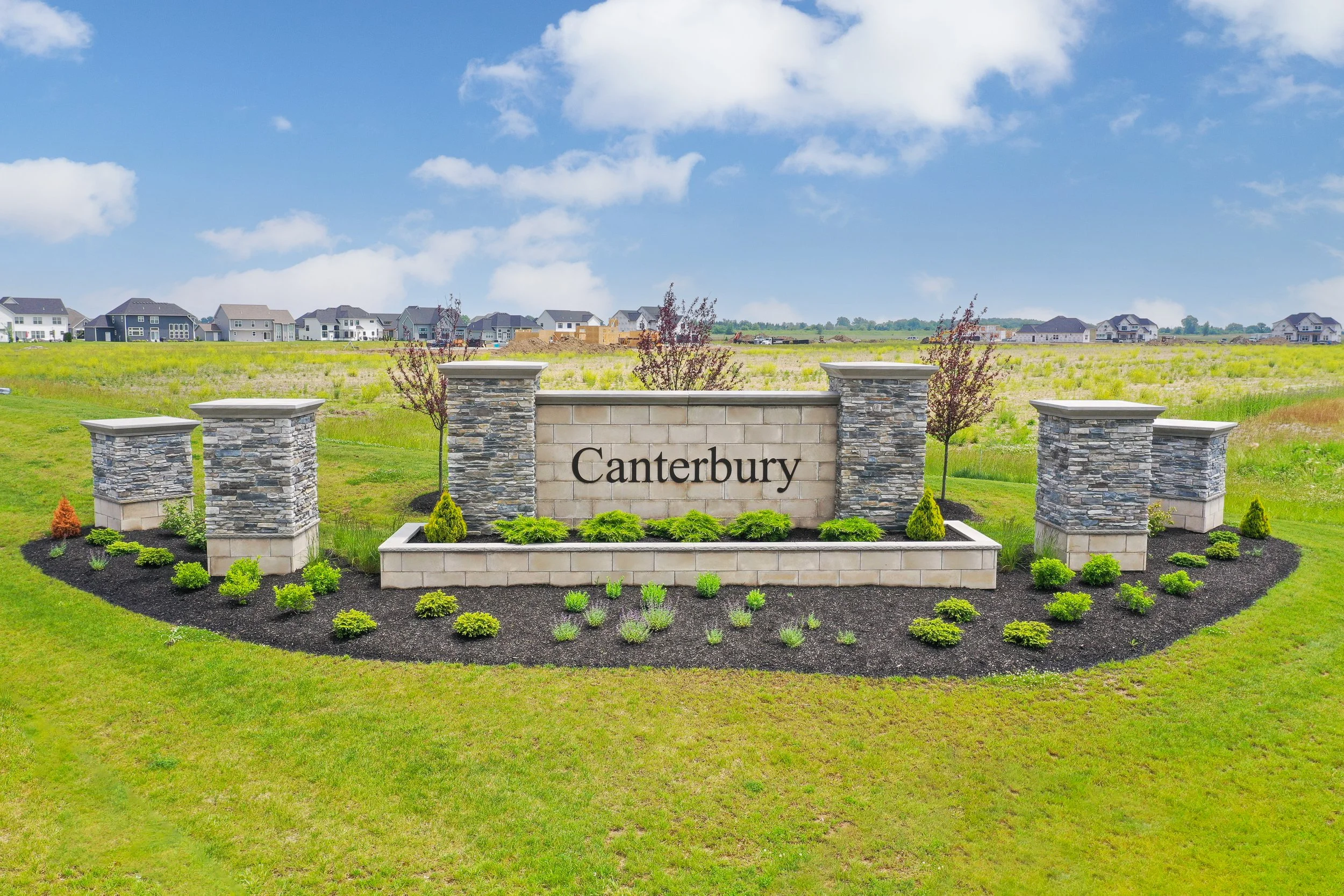 Sign that reads 'Canterbury' in front of a landscaped area with small green plants and young trees, with houses under construction across a grassy field in the background under a partly cloudy sky.