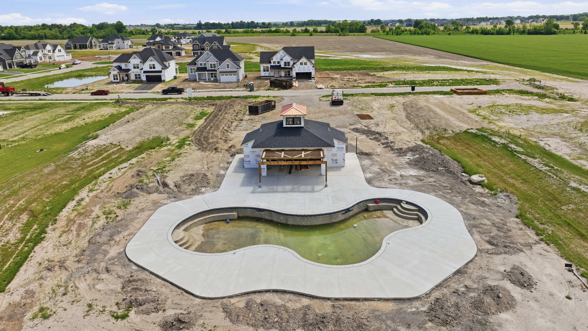Aerial view of a backyard with a kidney-shaped pool, empty and under construction, surrounded by a concrete patio, adjacent to a house under construction with visible framing and building wrap. In the background, there are multiple houses, a small po