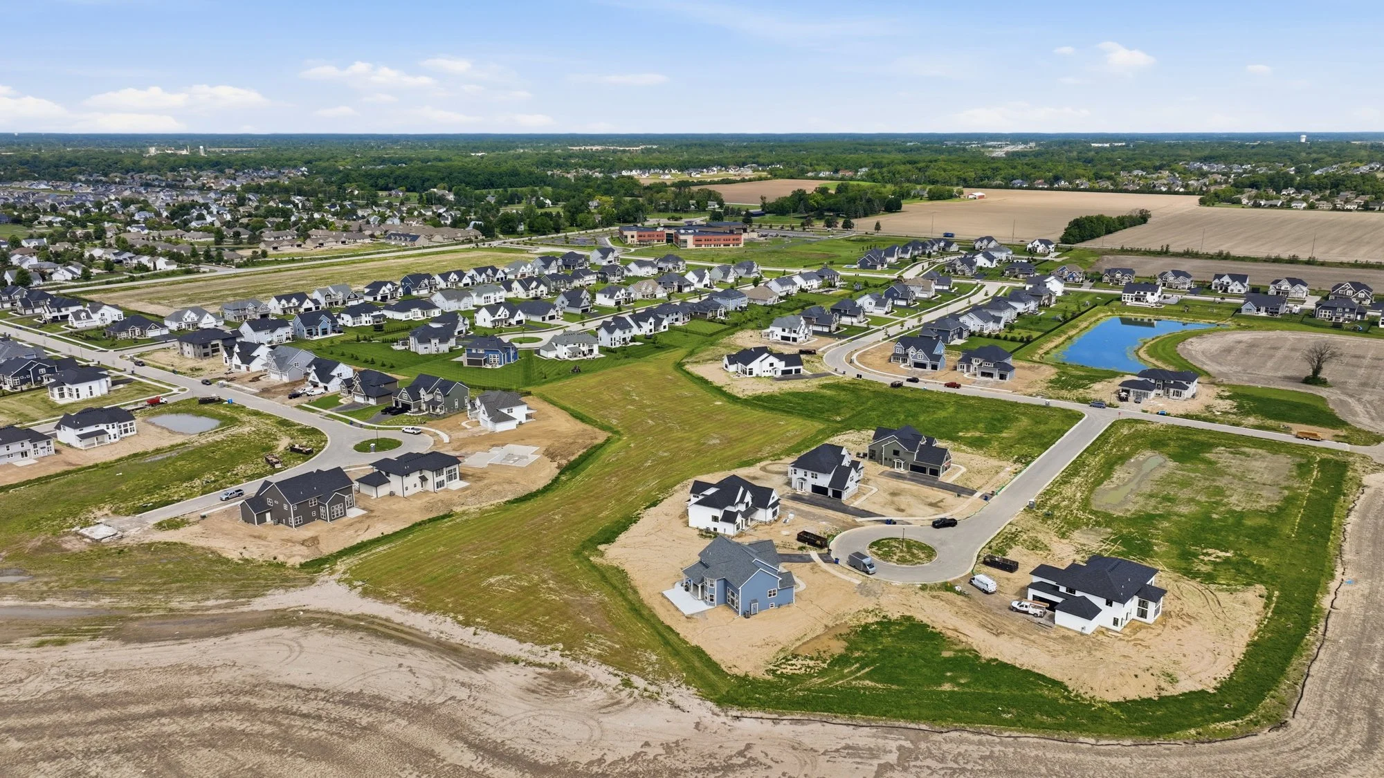 Aerial view of a suburban neighborhood under construction, with houses, roads, green lawns, and a small lake, surrounded by farmland and trees in the background.