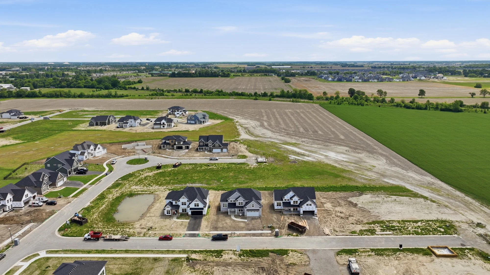 Aerial view of a residential construction site with several houses in various stages of completion, surrounded by open fields and farmland under a blue sky with some clouds.