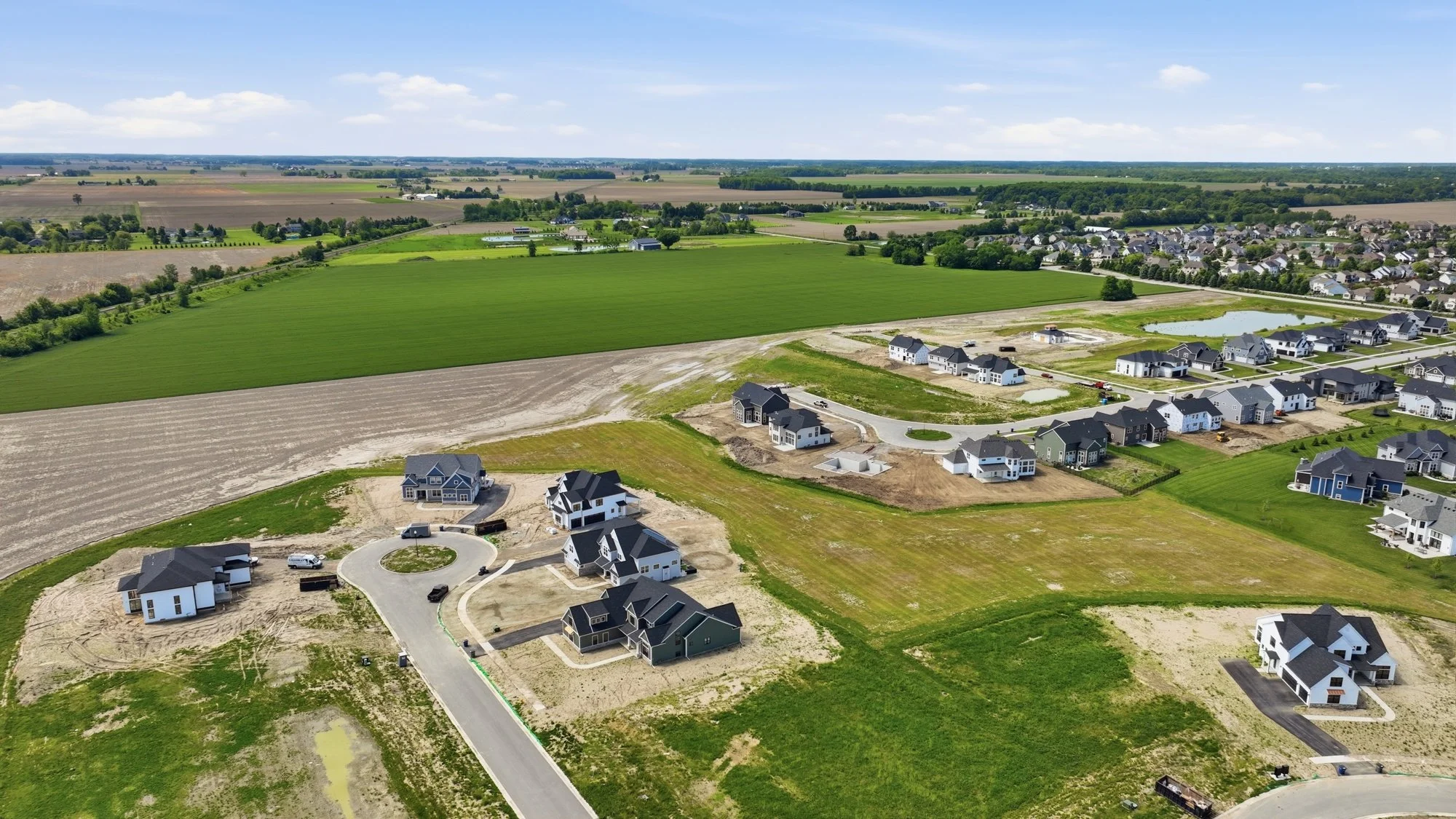 Aerial view of a suburban construction site with houses in various stages of completion, surrounded by green fields and farmland under a blue sky with scattered clouds.