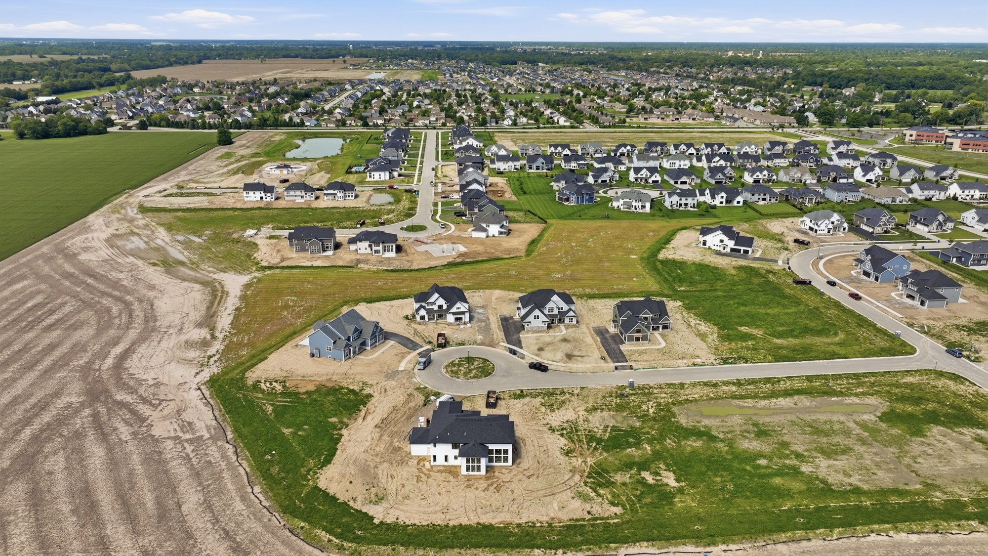 Aerial view of a new housing development with scattered houses under construction, roads, green lawns, and open land, near a large green field and a water body.