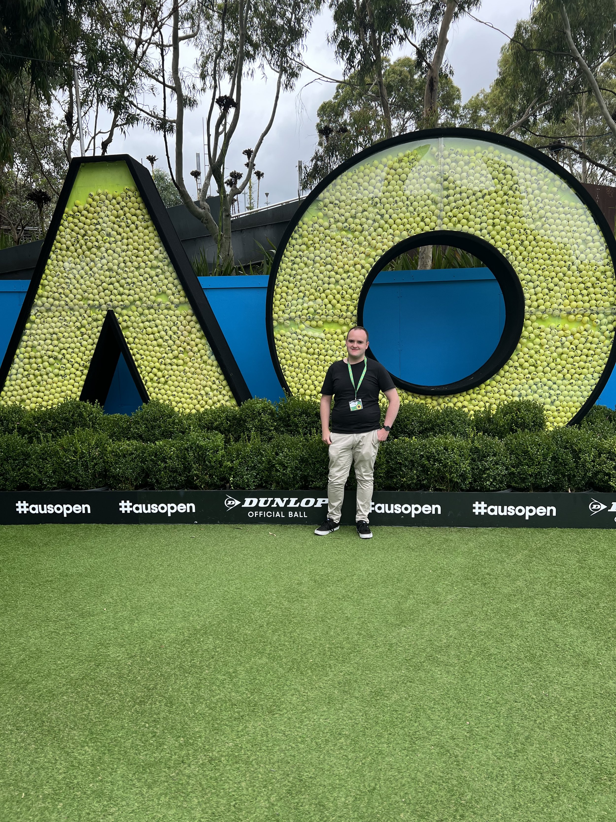   Image 1: Bryce standing in front of a giant Australian Open logo (AO) the Letters are filled with tennis balls. Bryce is wearing stone coloured long pant, a black t-shirt and a green lanyard with sunflowers on it.   