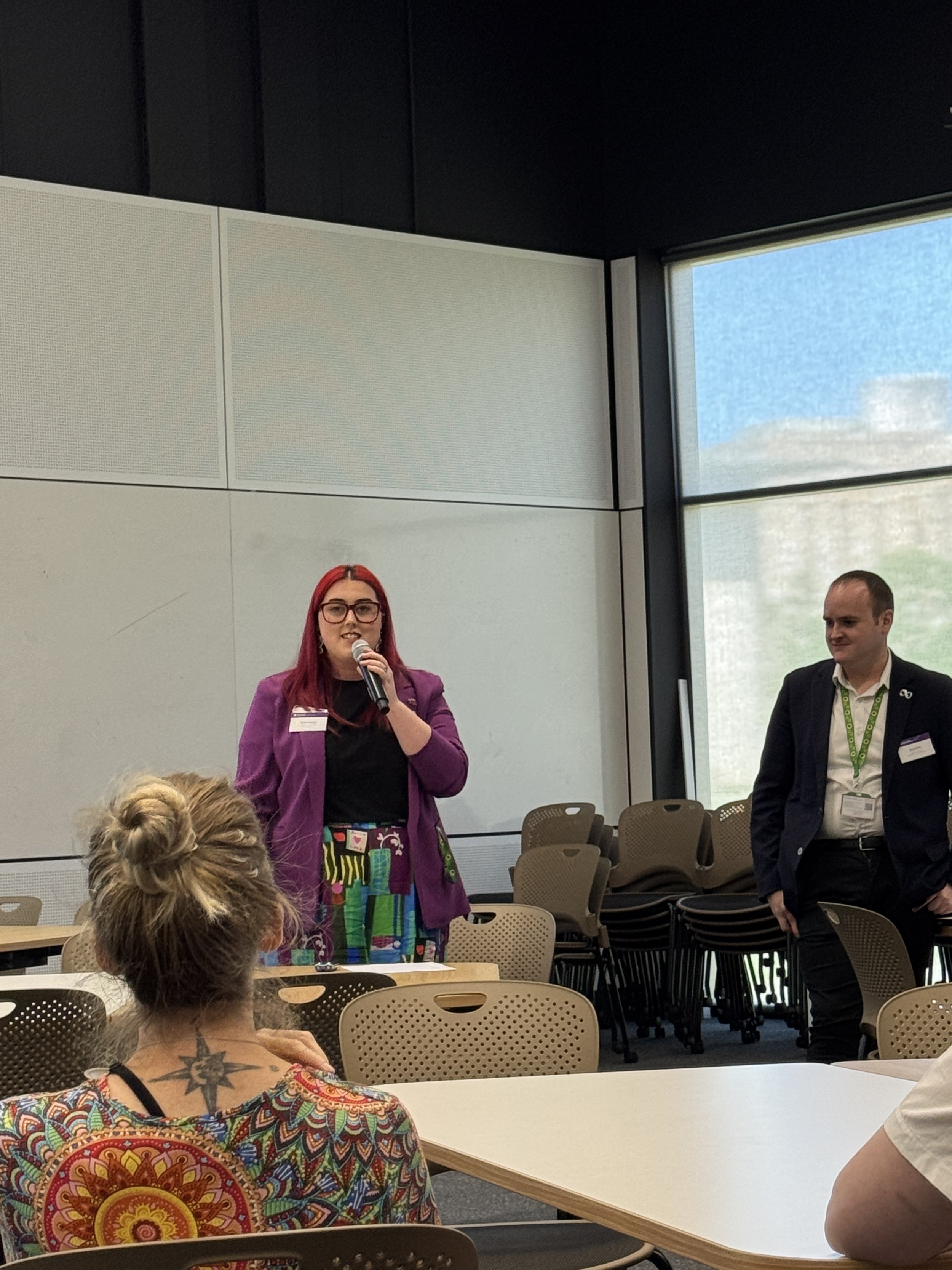          A photo of Tynah Pearson from Victoria University presenting, Tynah is a female in her 20s with long red hair, glasses and wearing a purple blazer, black top and colourful pants. Bryce can be seen standing beside Tynah, ready to present. Bry