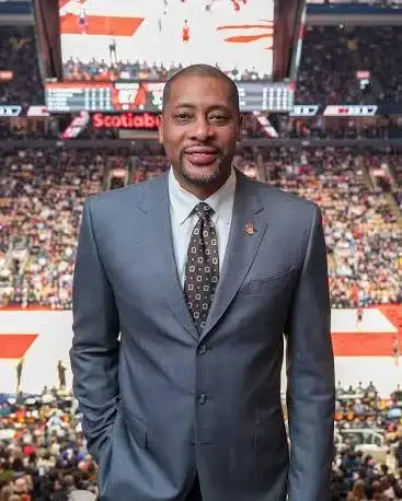 A man in a gray suit and tie standing in a sports arena with a large crowd and scoreboard in the background.