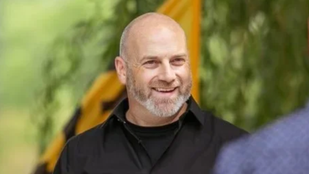 A smiling man with a shaved head and beard, wearing a black shirt, outdoors with blurred greenery in the background.