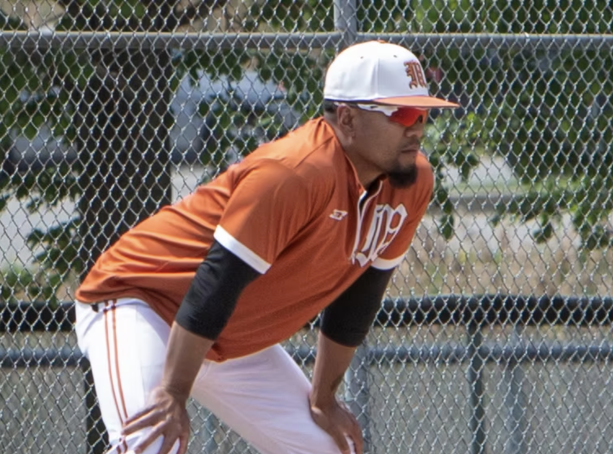 A baseball player wearing sunglasses, a white cap, orange jersey, and white pants, crouched behind a chain-link fence on the field.