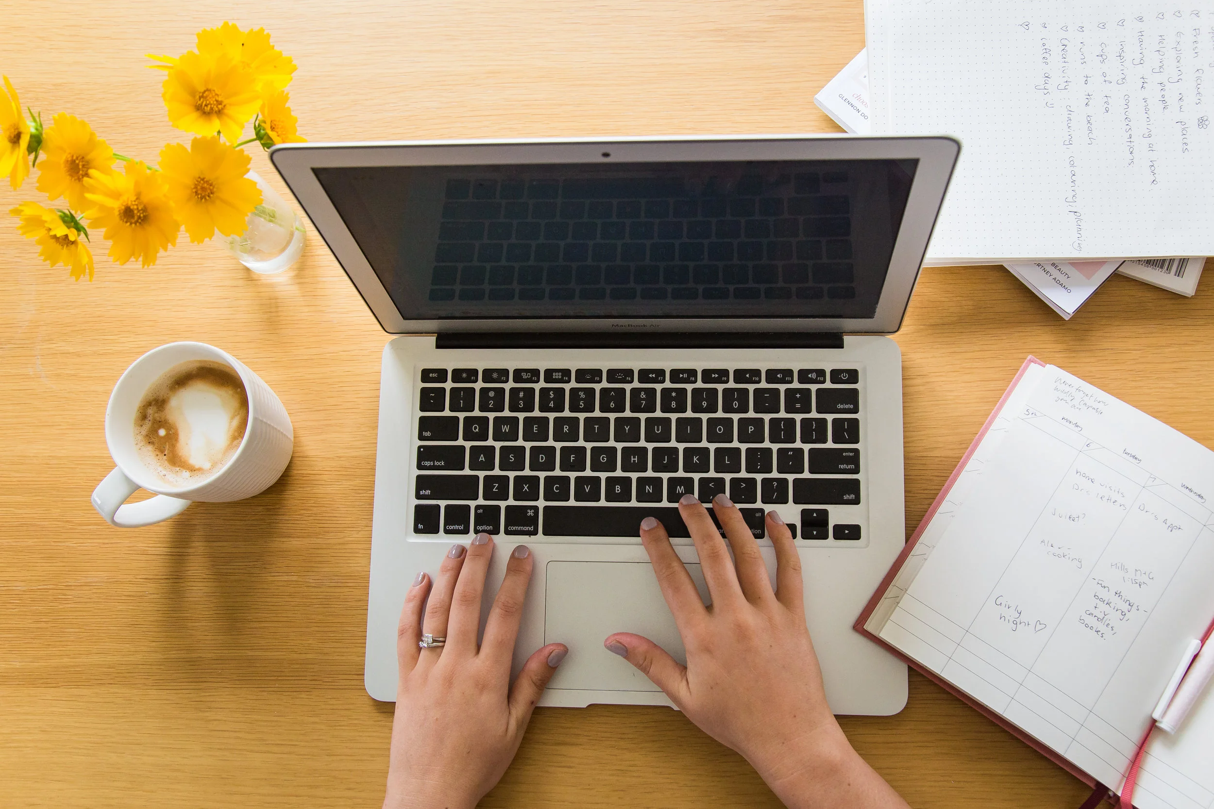 Person typing on a laptop with a notebook, yellow flowers, and a coffee cup on a wooden desk.