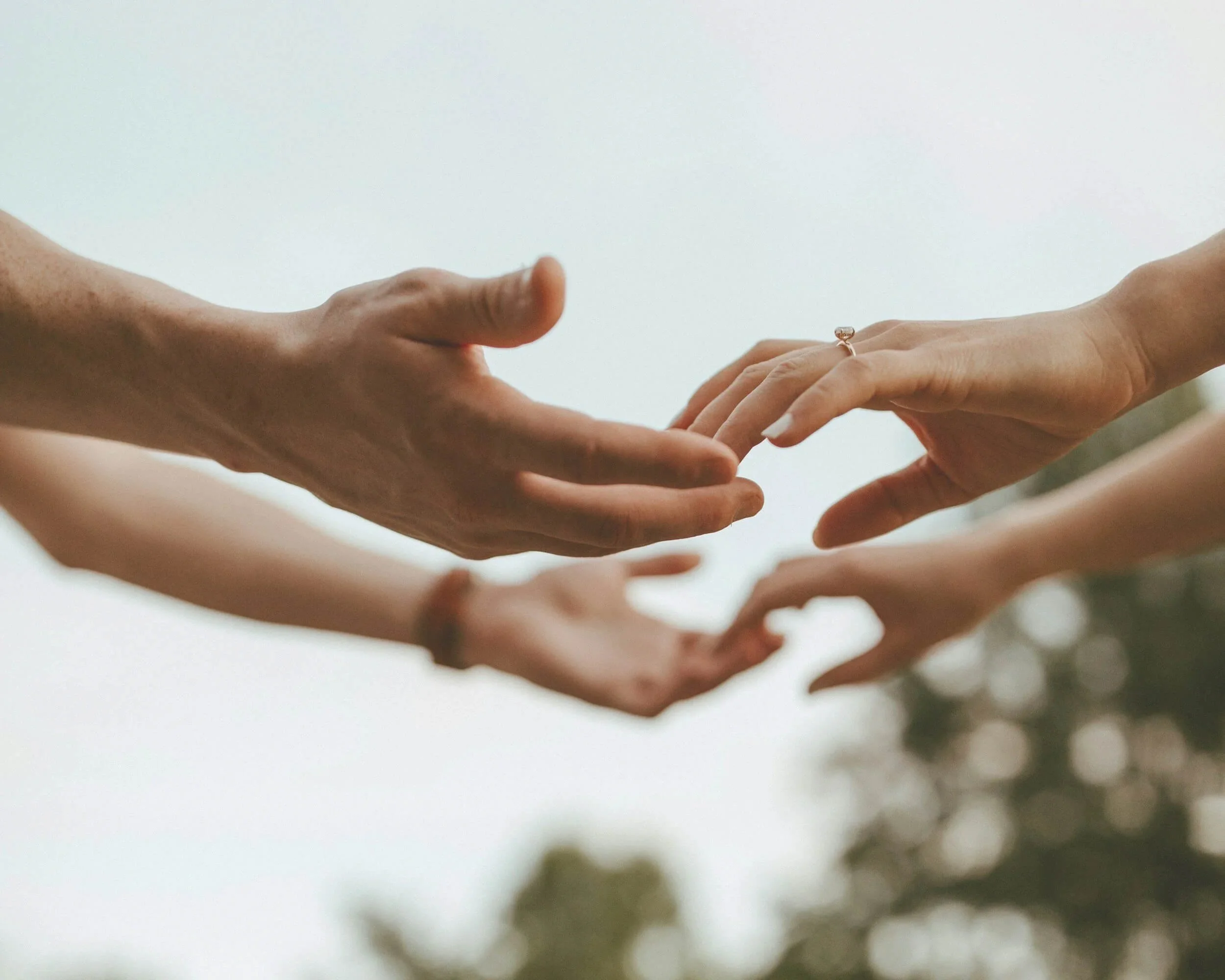 Four hands reaching towards each other against a sky background, with one hand wearing a ring.
