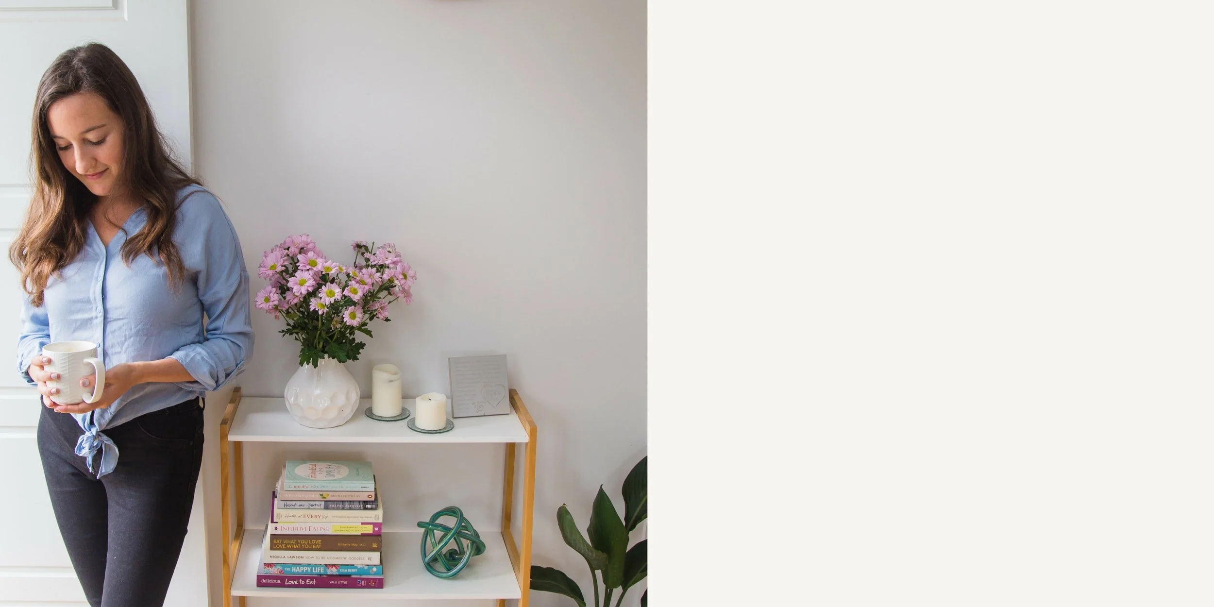 Woman holding a mug next to a white shelf with pink flowers, candles, books, and decorative items.