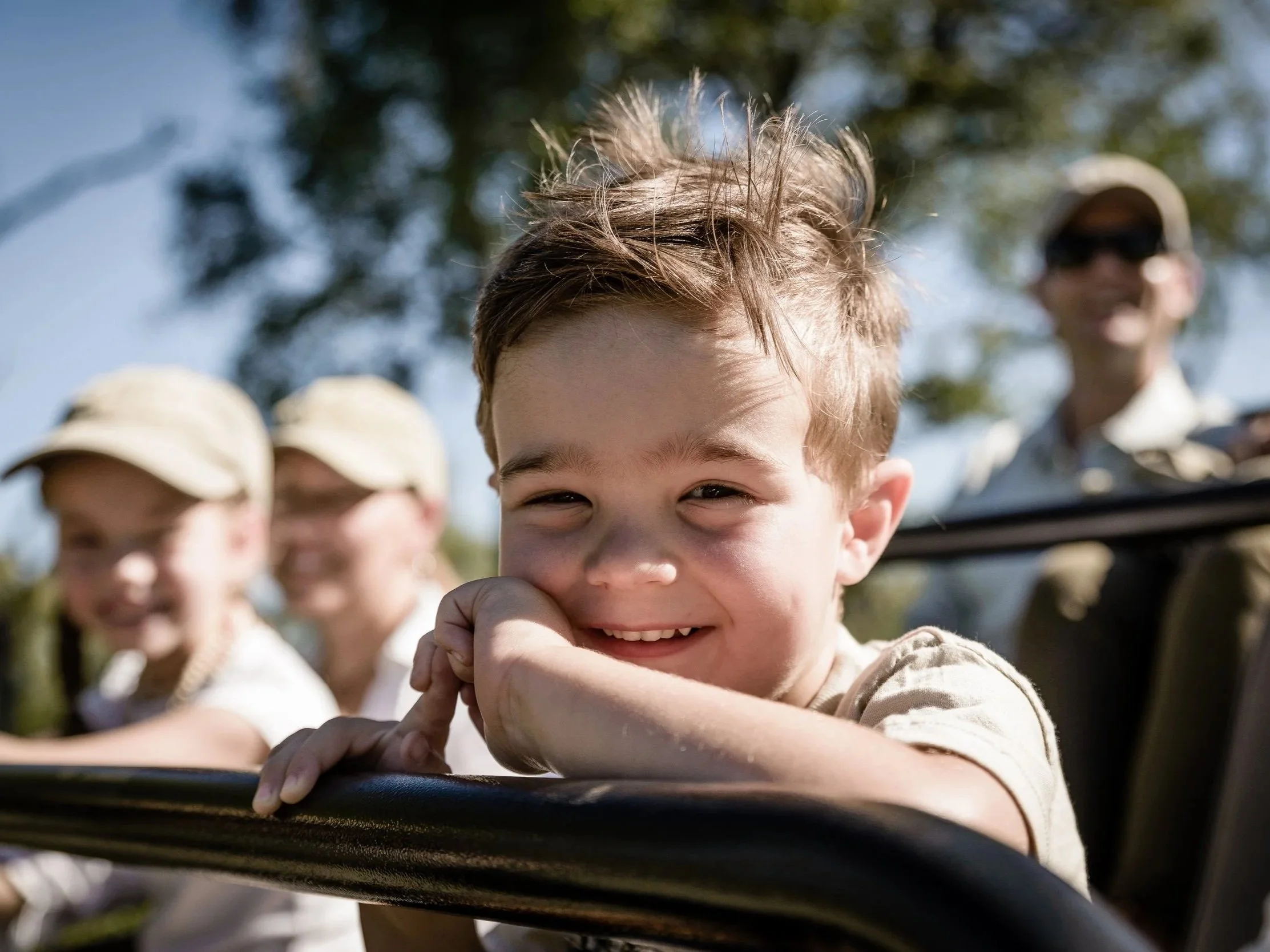 Close-up of a smiling boy with brown hair riding in a vehicle with other children and adults outside during daytime.