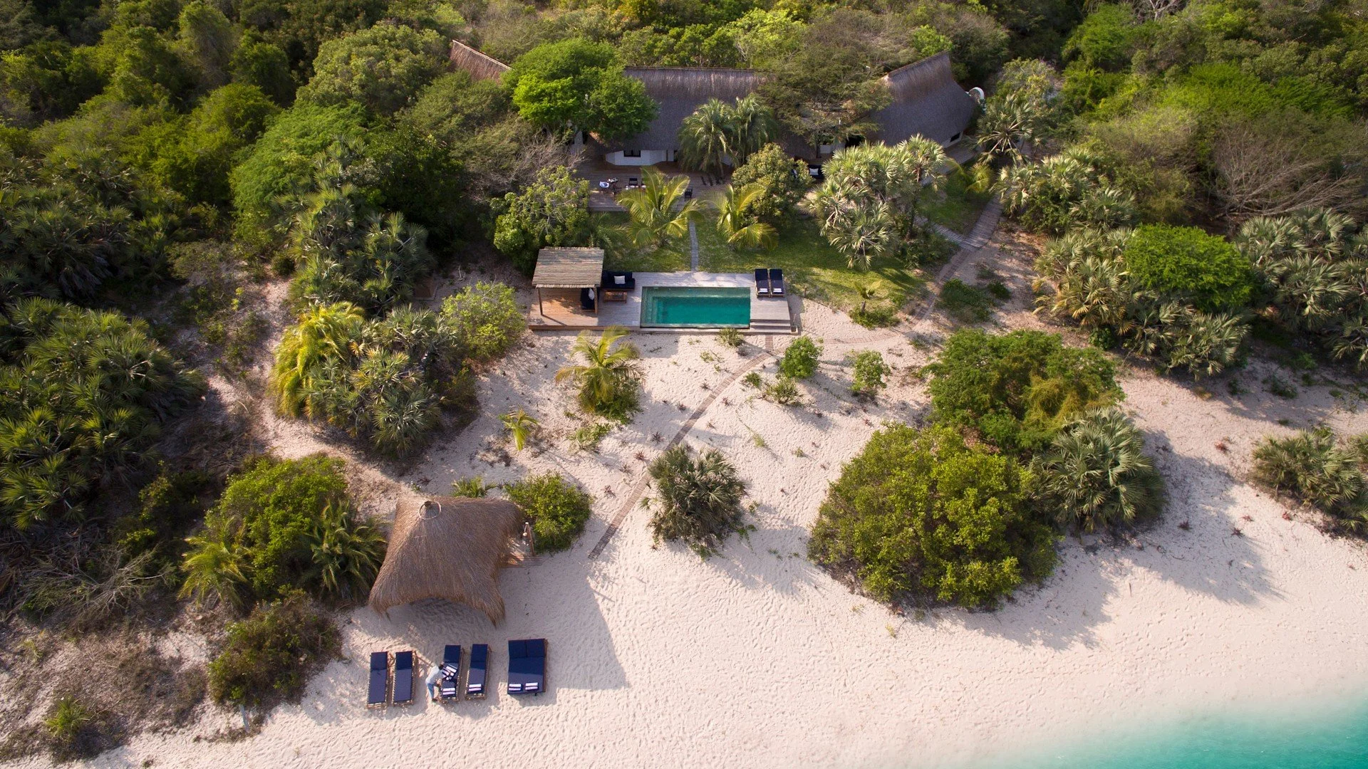 An aerial view of a beachside property surrounded by lush green trees, featuring a swimming pool, a shaded outdoor lounge area, a sandy beach with lounge chairs, and a thatched-roof hut, with turquoise water at the shoreline.