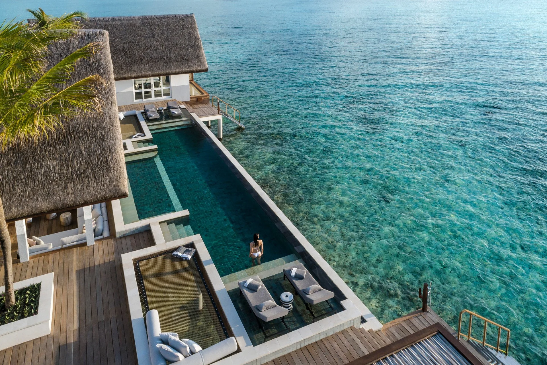Maldives Overwater bungalow with private pool overlooking clear ocean water, a woman in a swimsuit stands in the pool, and a woman lounges nearby on a deck chair.