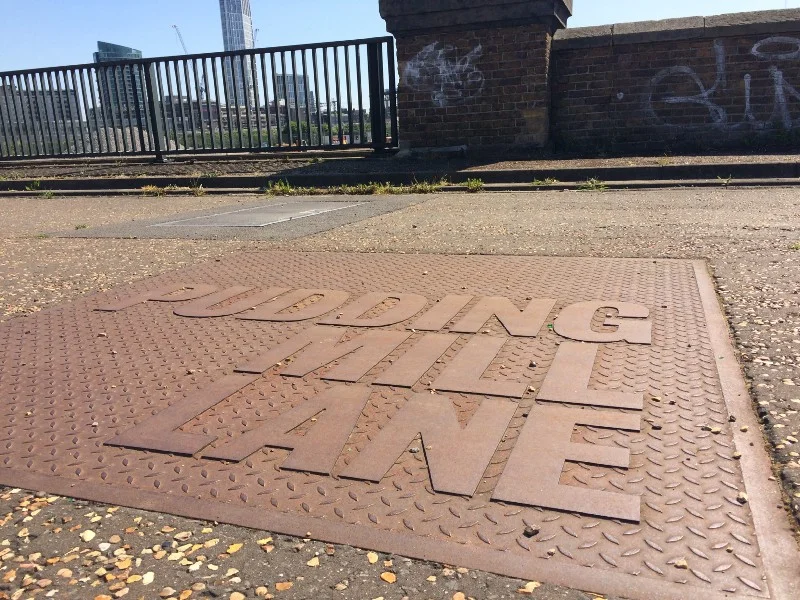 Stylised utility cover on the Greenway. Through the fence, one of the rubble piles.